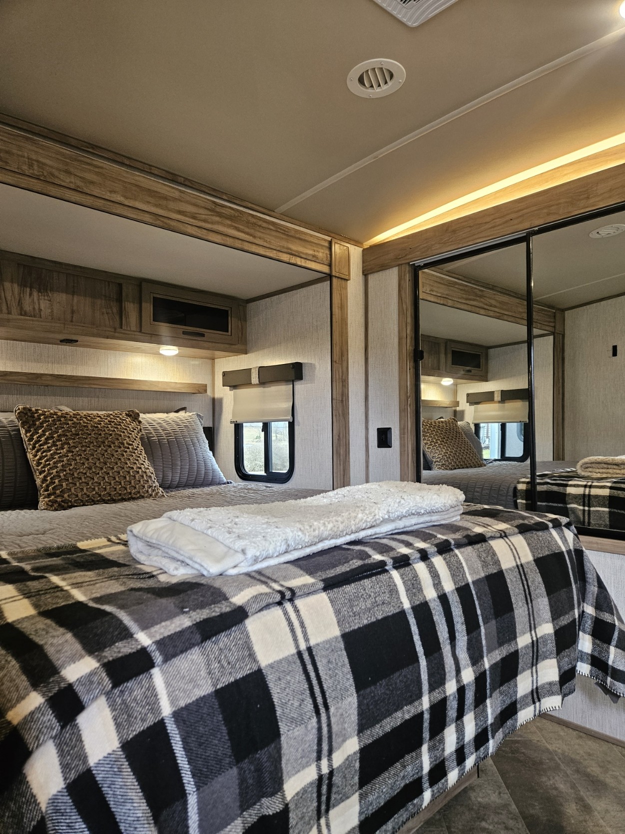 Cozy RV rental bedroom interior, featuring a bed with plaid blanket, warm lighting, wooden accents, & mirrored closet.. Forest River Palomino Solaire 2021