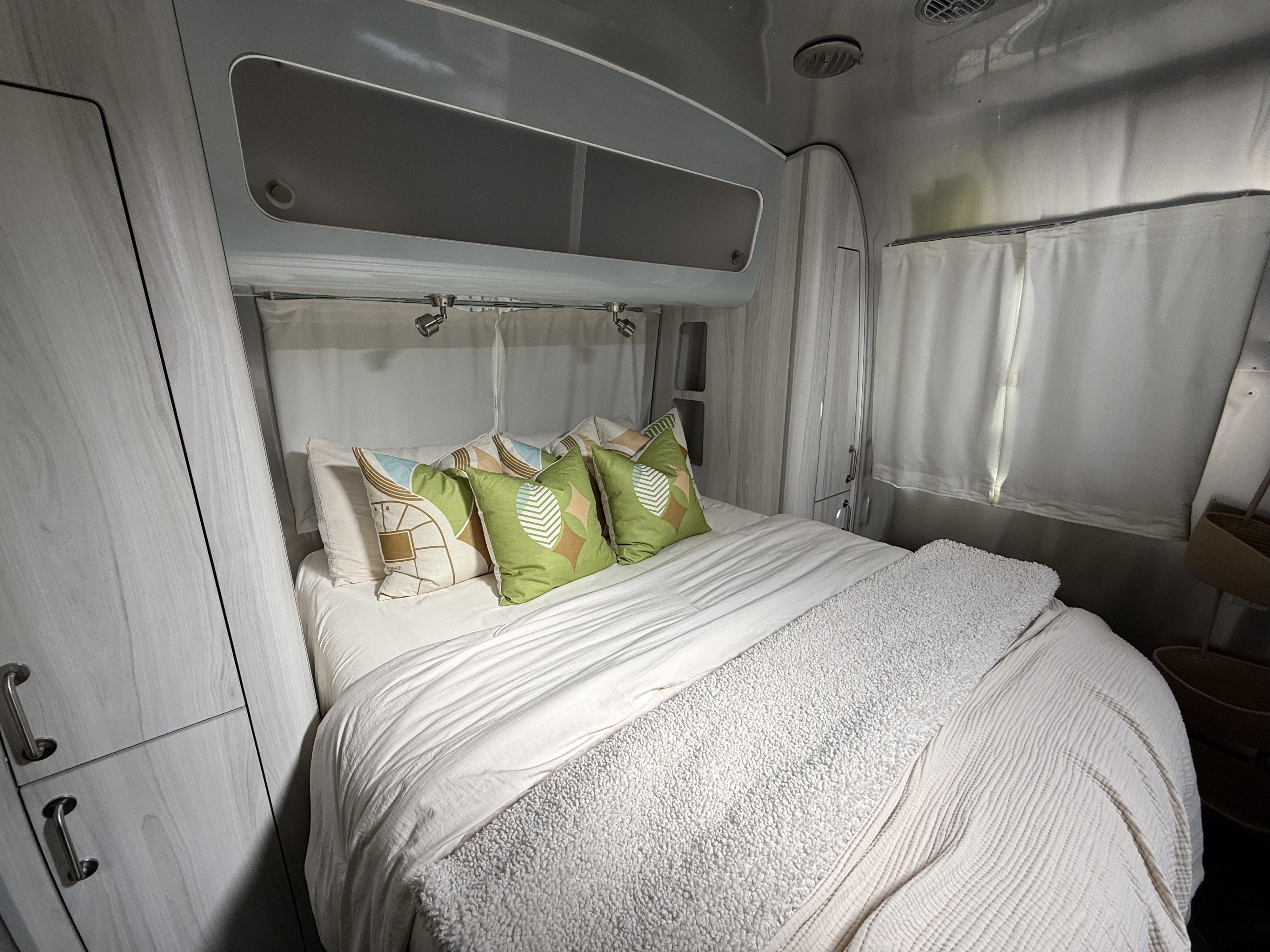 This RV rental's cozy bedroom features a comfortable bed with colorful pillows, stylish cabinets, and overhead lighting for travel.. Airstream International 2021
