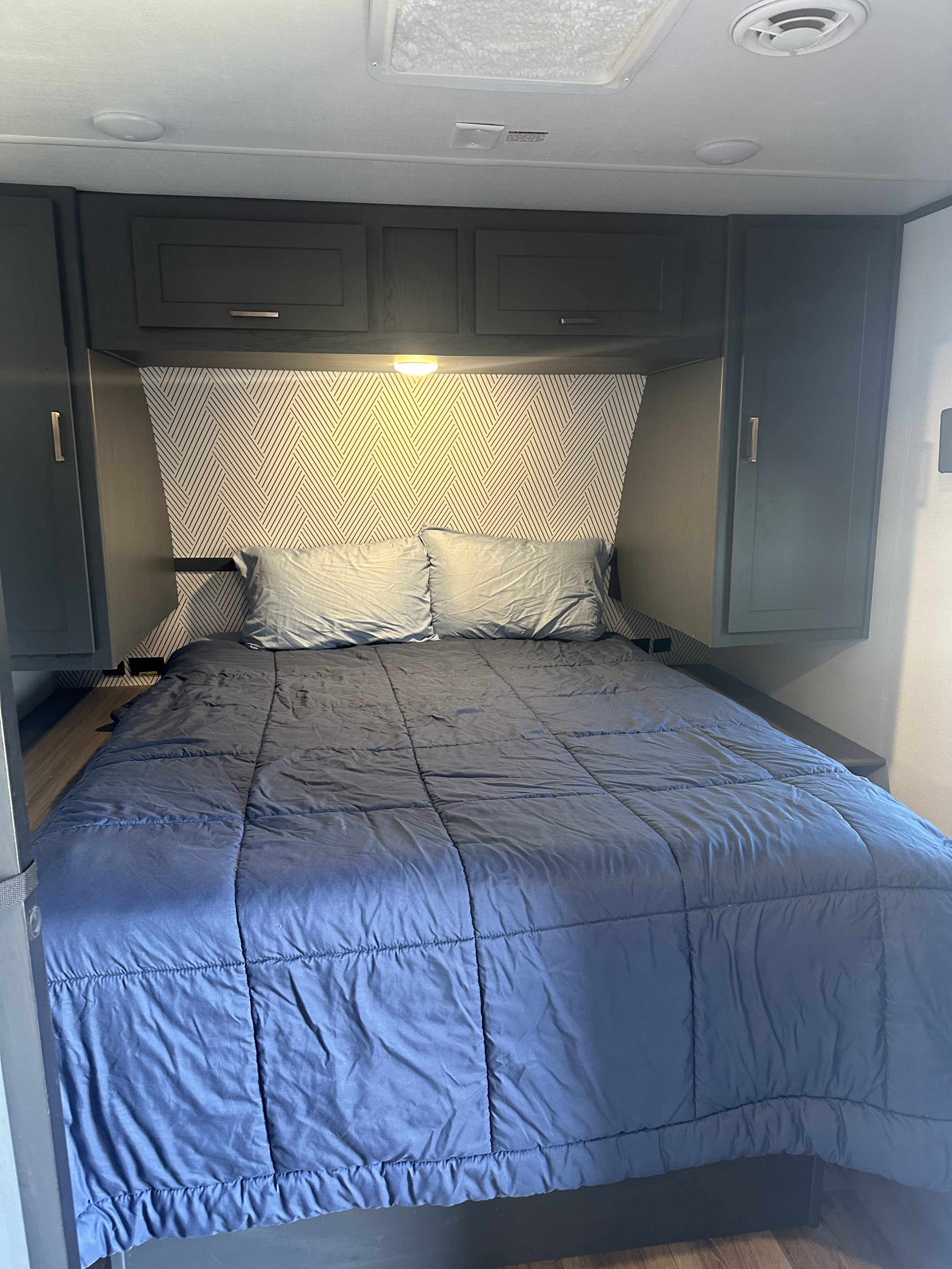 RV rental bedroom featuring a blue bed with pillows, geometric headboard wallpaper, overhead cabinets, and ceiling fixtures.. Keystone RV Bullet Crossfire 2024