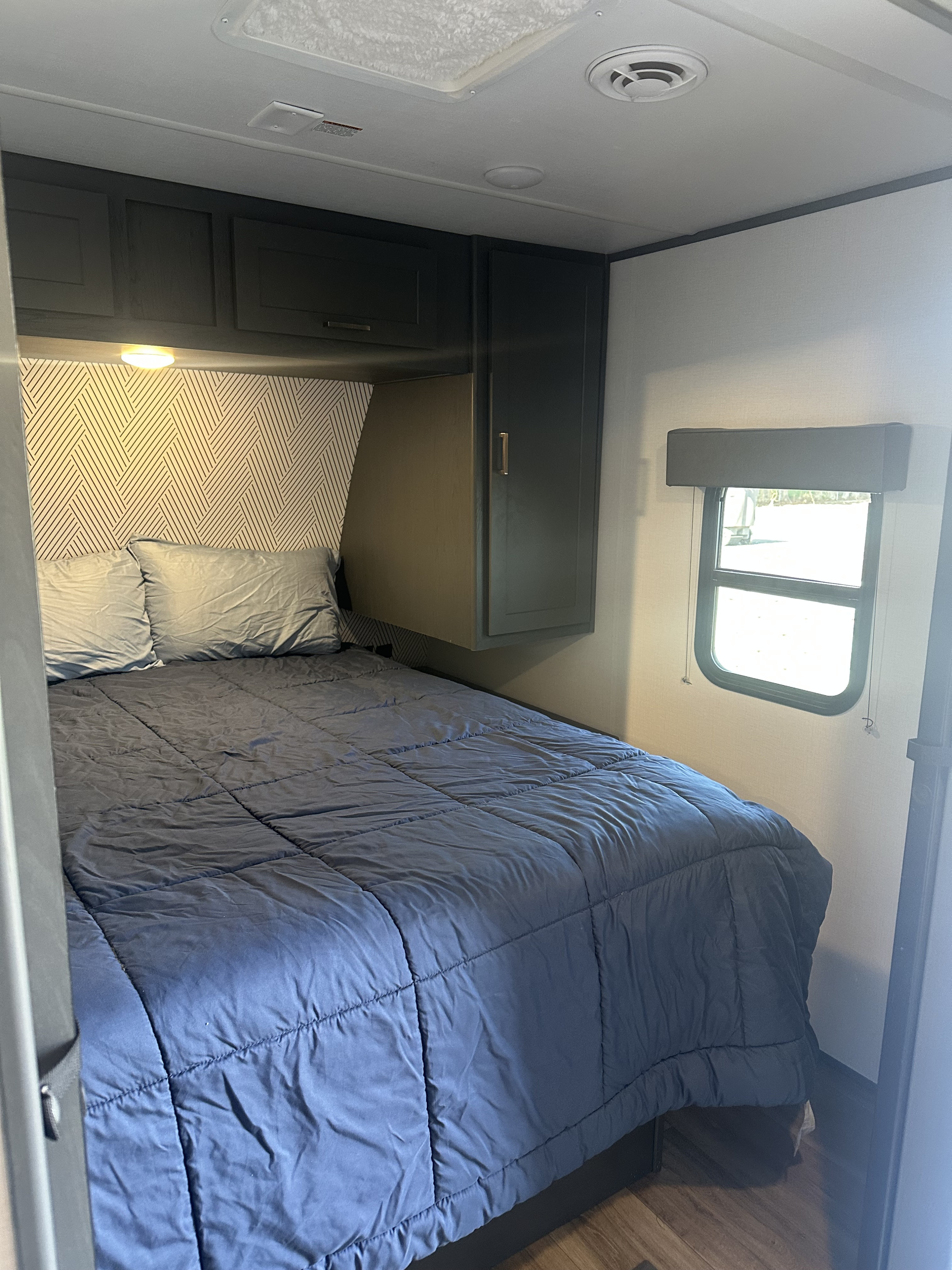 RV rental bedroom featuring dark cabinetry, geometric wallpaper, and a cozy blue bed and pillows with an exterior window.. Keystone RV Bullet Crossfire 2024