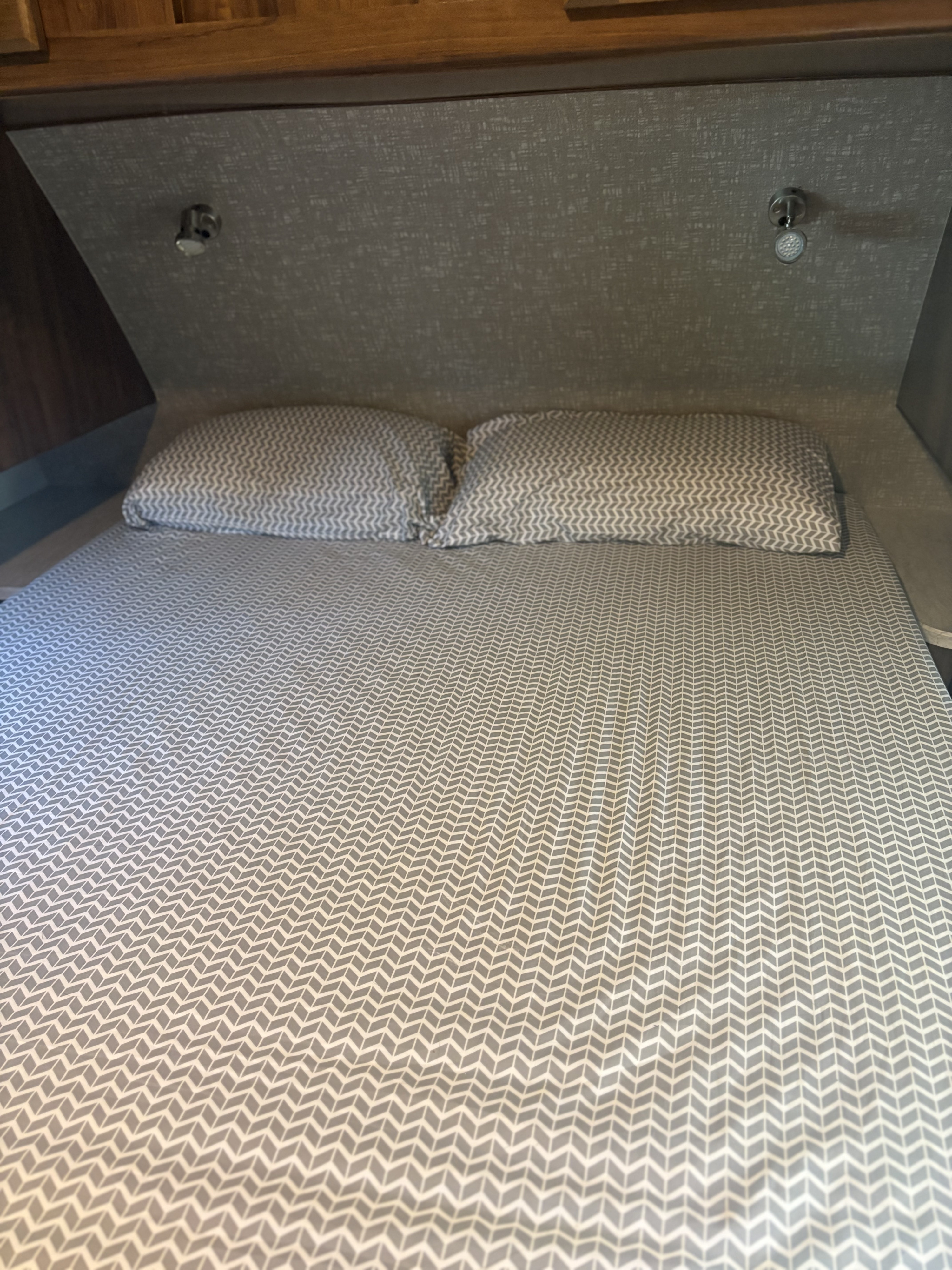 This cozy RV rental bed features gray chevron bedding, two pillows, and overhead reading lights, ideal for travel.. Heartland RVs Mallard 2019
