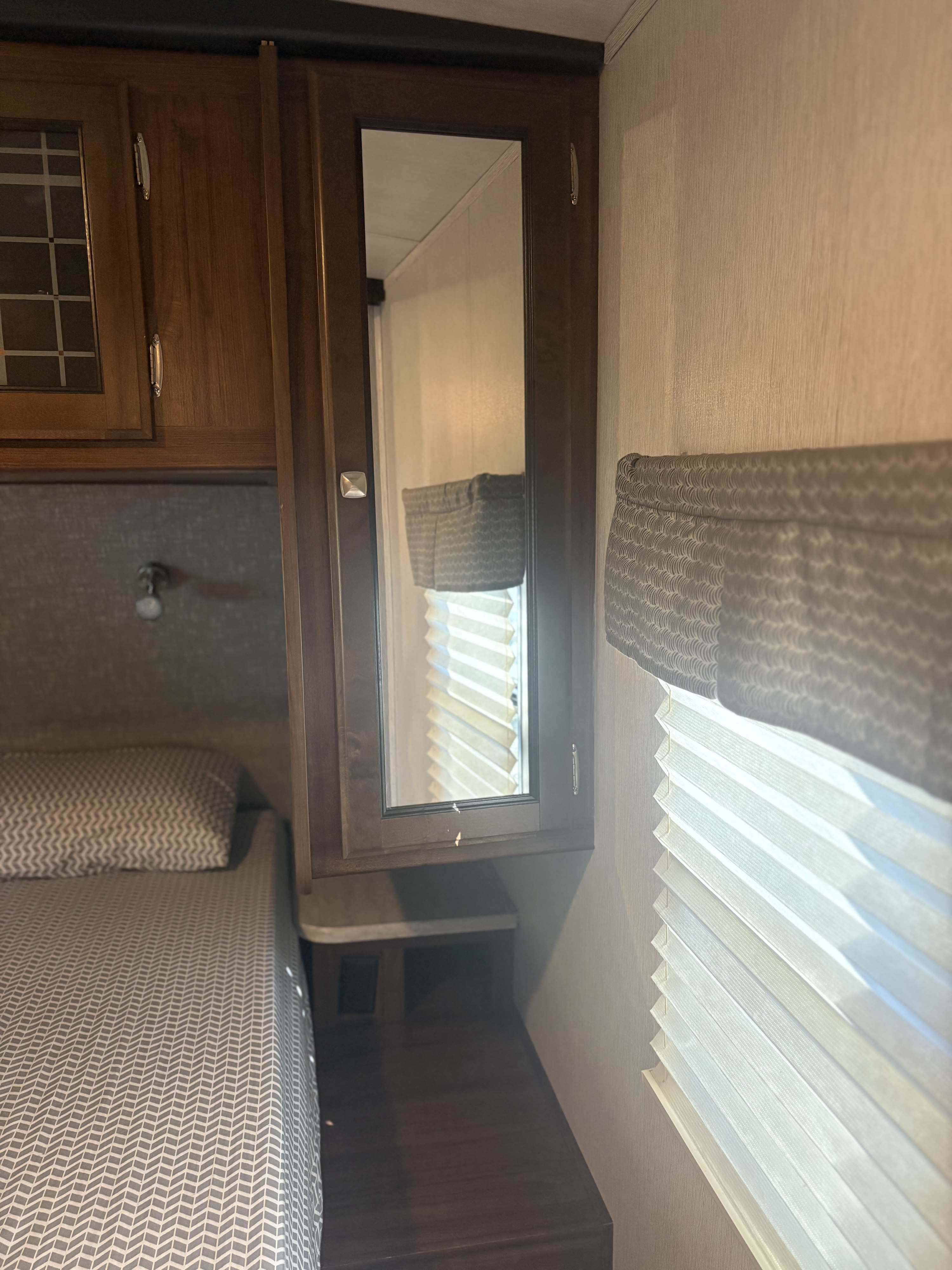 Cozy RV rental bedroom featuring patterned bedding, wooden cabinets, a mirror, and a window with cellular blinds.. Heartland RVs Mallard 2019