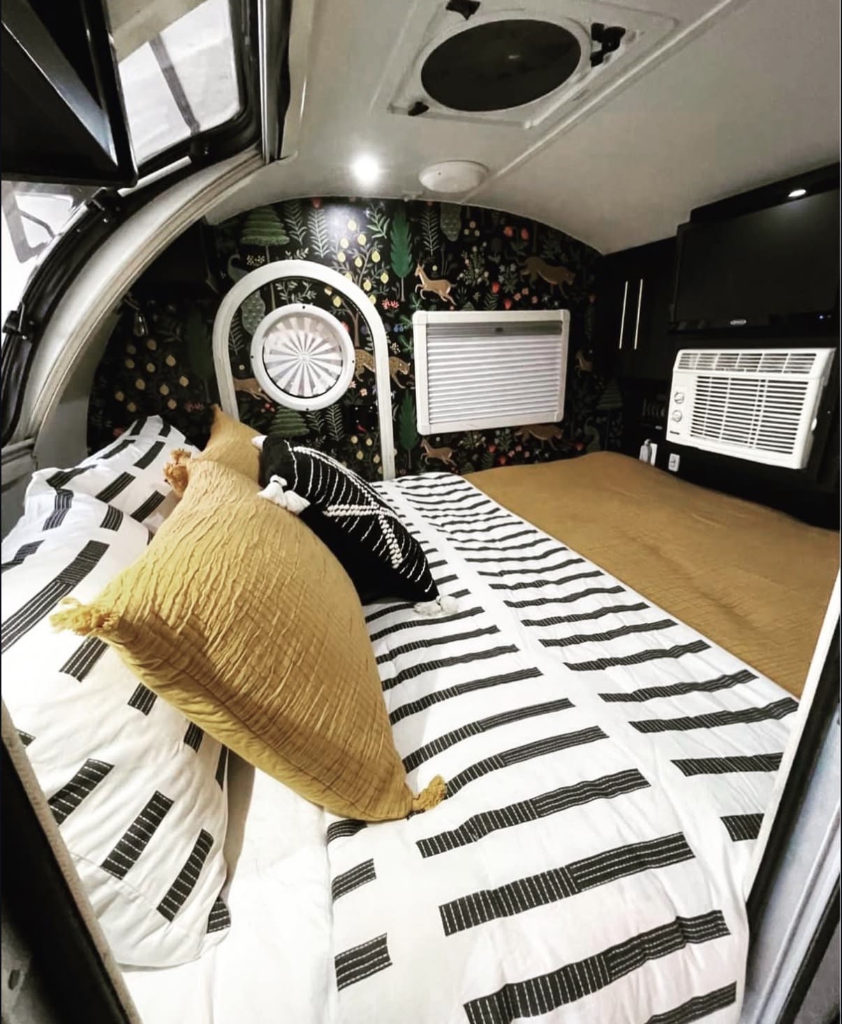 Cozy rv rental bedroom with a striped bedspread, yellow pillow, dark forest-themed wallpaper, and AC unit.. nuCamp T@G 2017
