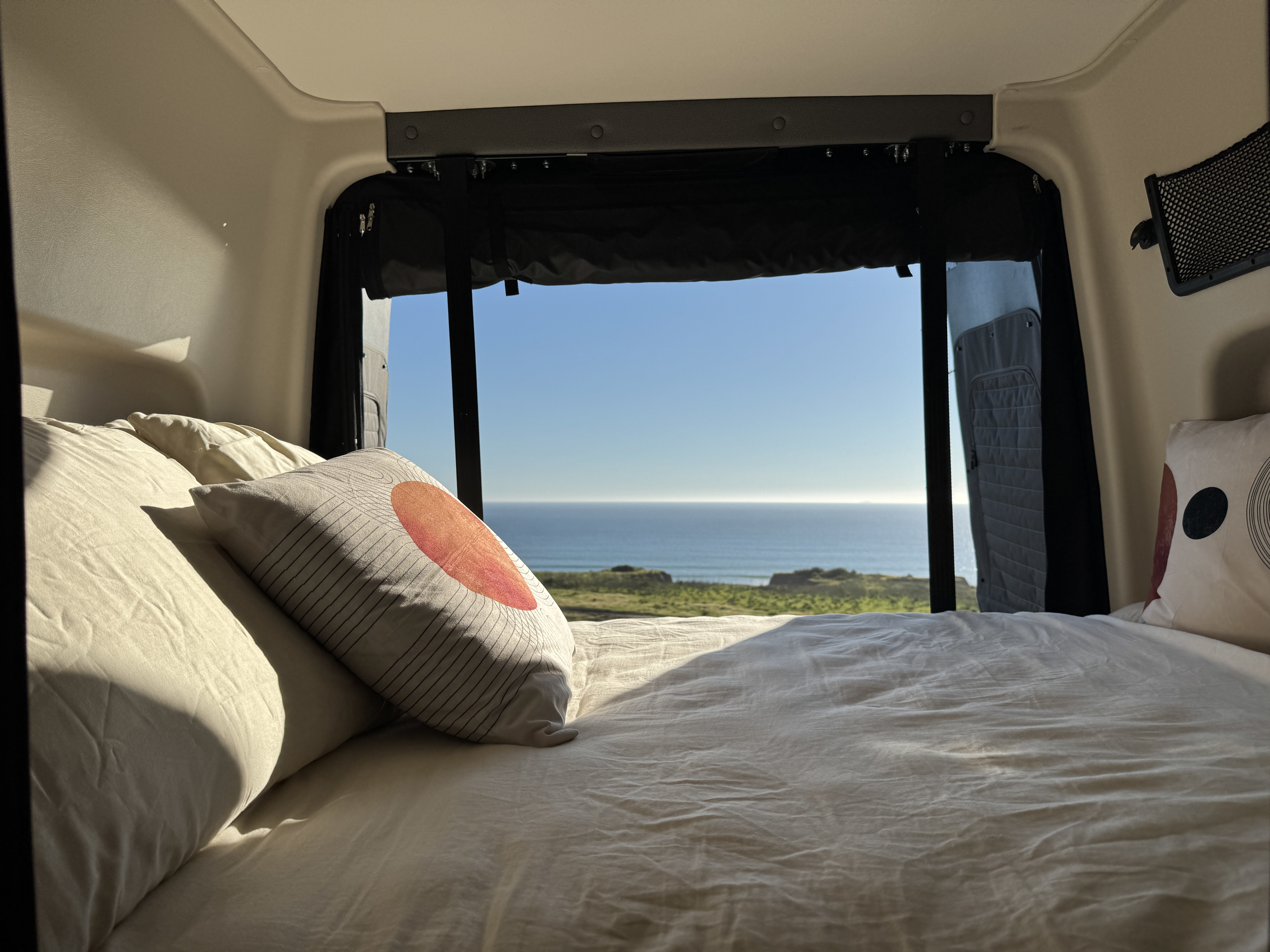 RV rental bed with a scenic ocean view from the camper's back. Cozy interior showing bedding and coastal landscape outside.. Winnebago Revel 2025