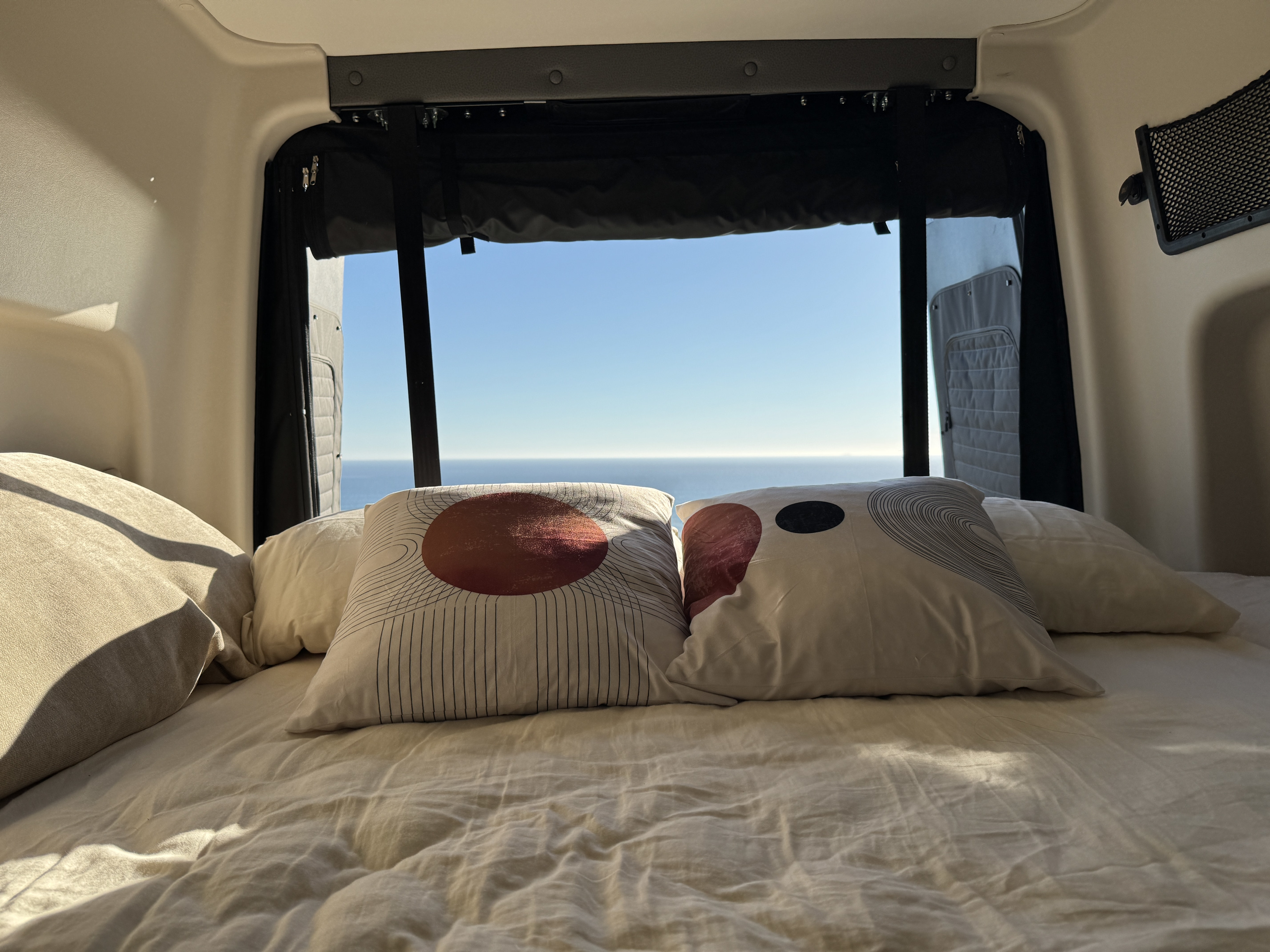 This rv rental features a cozy bed with an expansive ocean view from the rear window. Pillows and bedding are visible in sunlight.. Winnebago Revel 2025