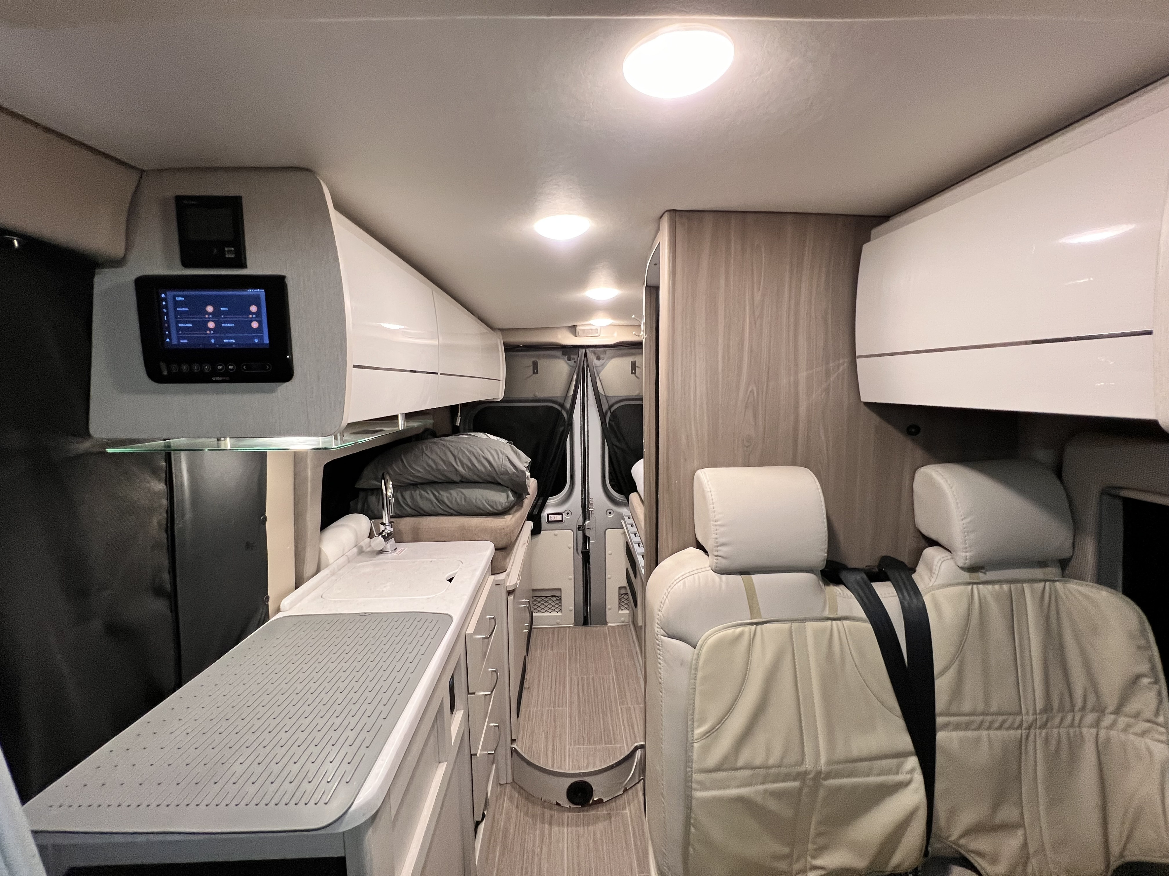 Inside an RV rental, a compact living space features a kitchen, bed area, passenger seats, and a wall control panel.. Thor Rize 2022