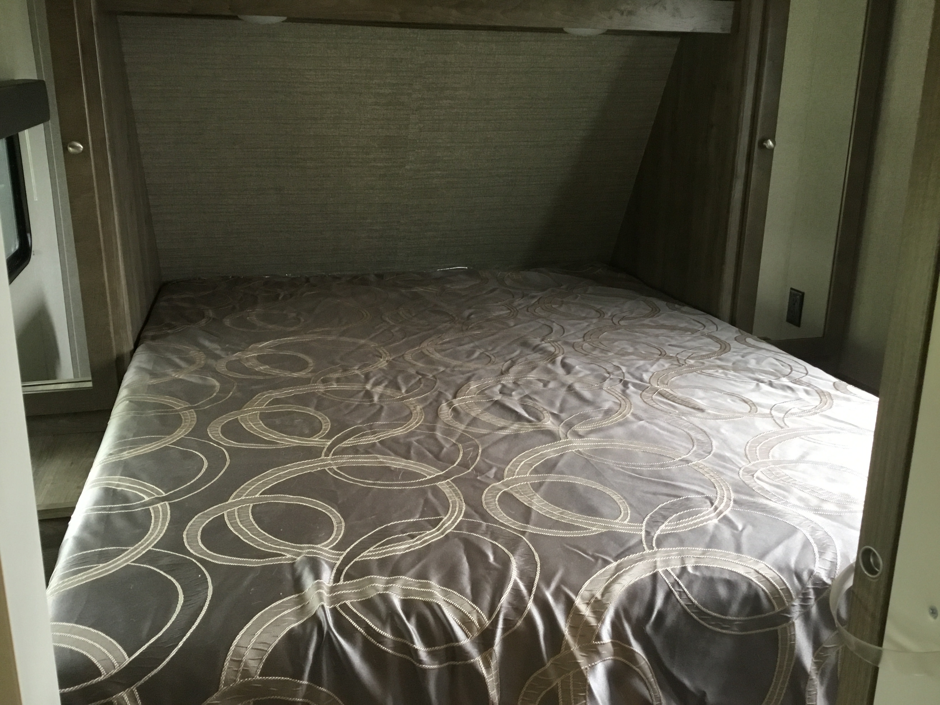 This rv rental showcases a small bedroom with a gray patterned bed, soft overhead lights, and a side window, ideal for travel.. Dutchmen Aspen Trail 2019