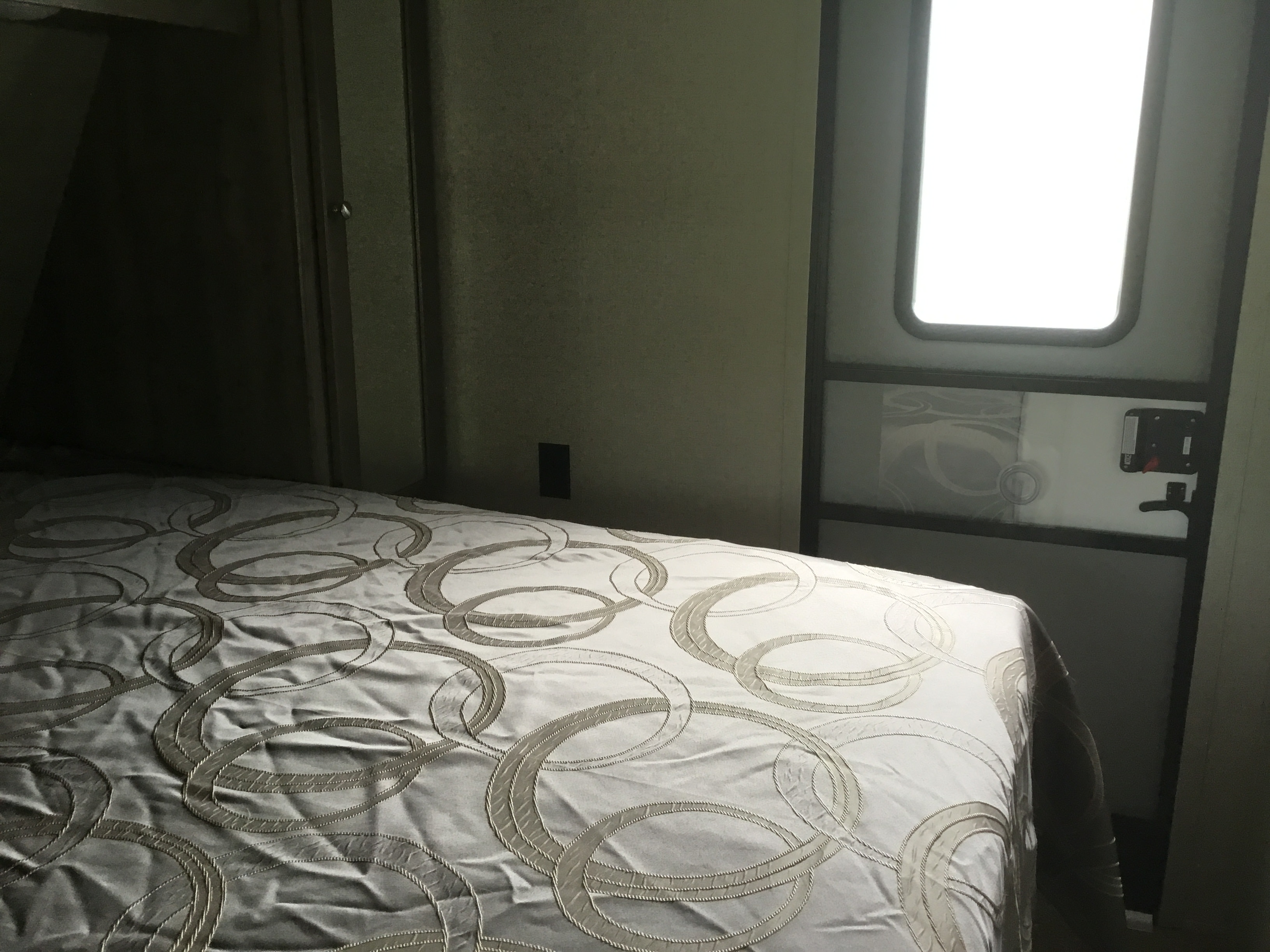 RV rental bedroom with a patterned comforter on the bed, and a door featuring a bright window and latch.. Dutchmen Aspen Trail 2019