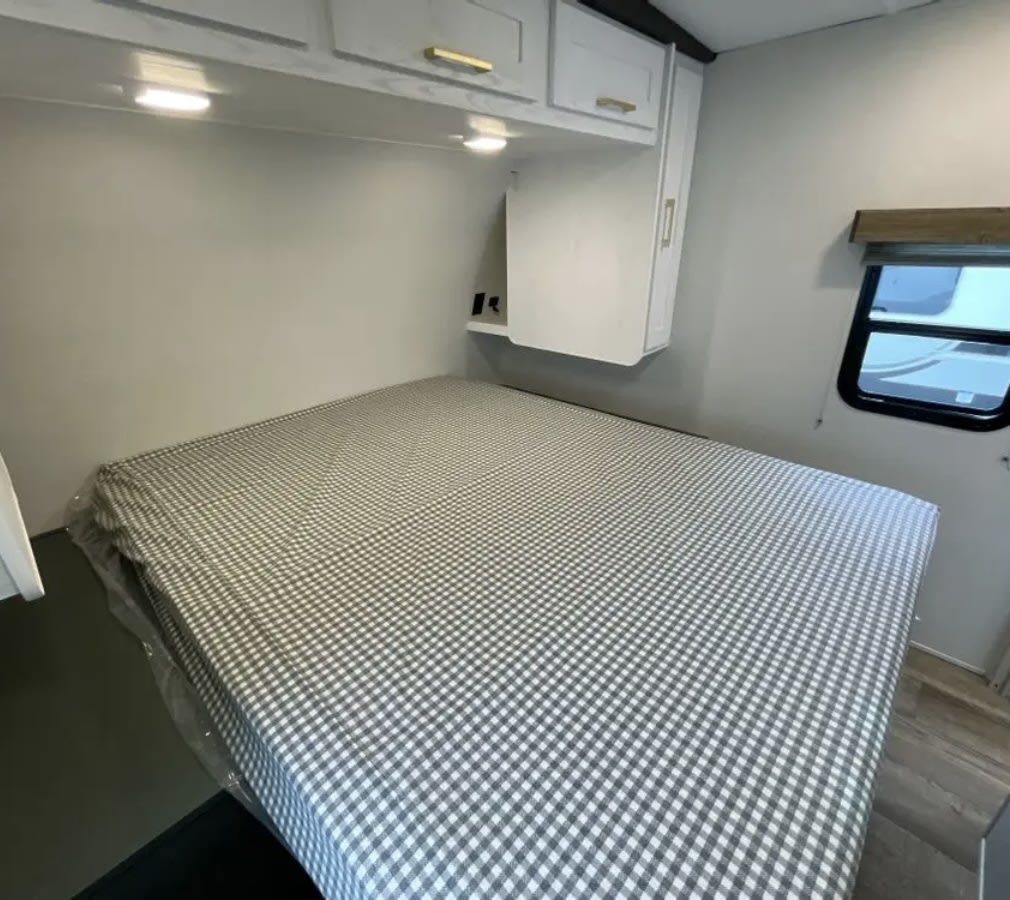 Cozy RV rental bedroom featuring a checkered bed, bright overhead lights, white cabinets, and a side window.. Heartland RVs Eddie Bauer 2024