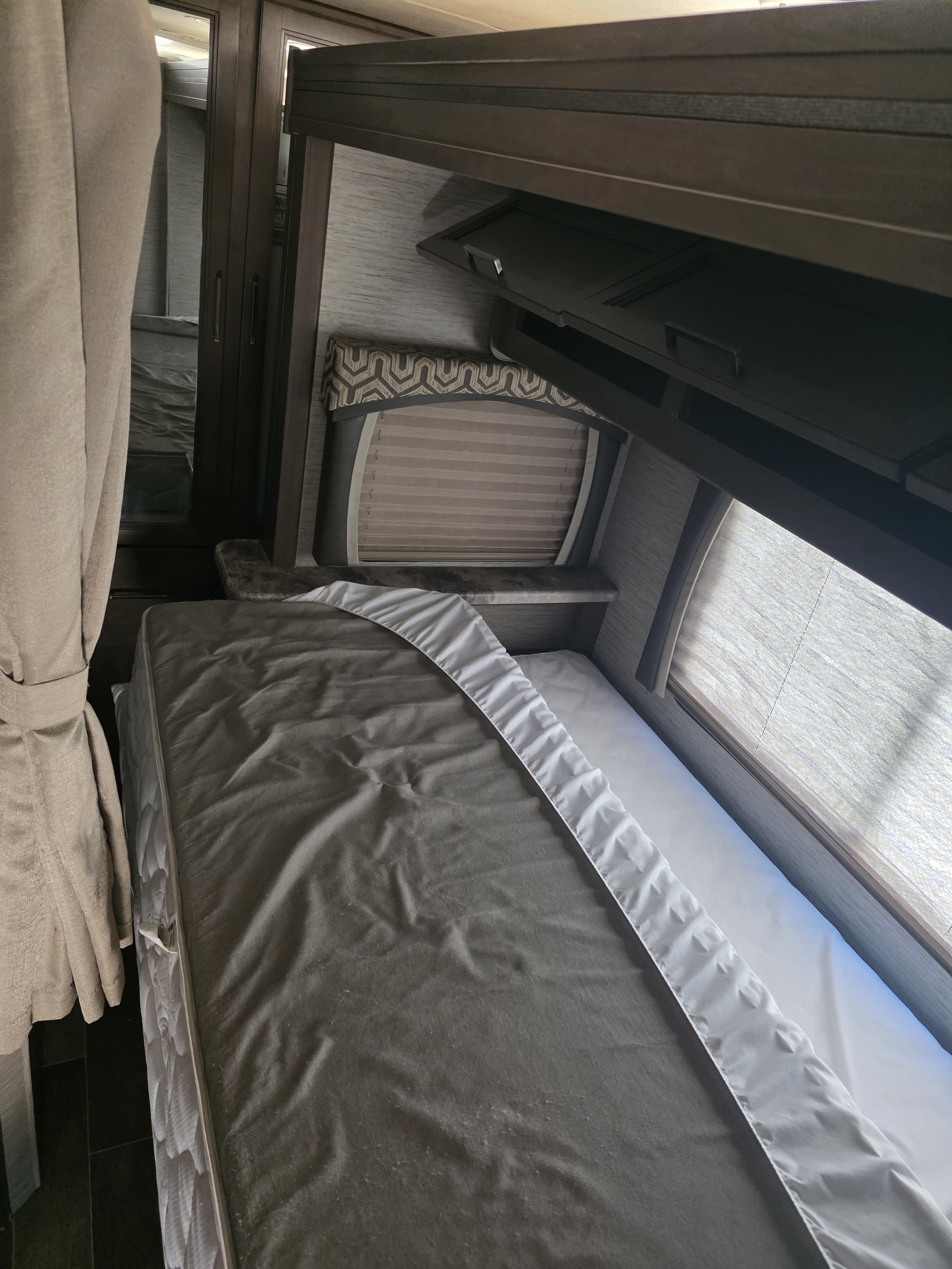 RV rental sleeping area: bunk bed with dark mattress, white sheet, window with blinds, and wood cabinetry.. Thor Chateau 2021