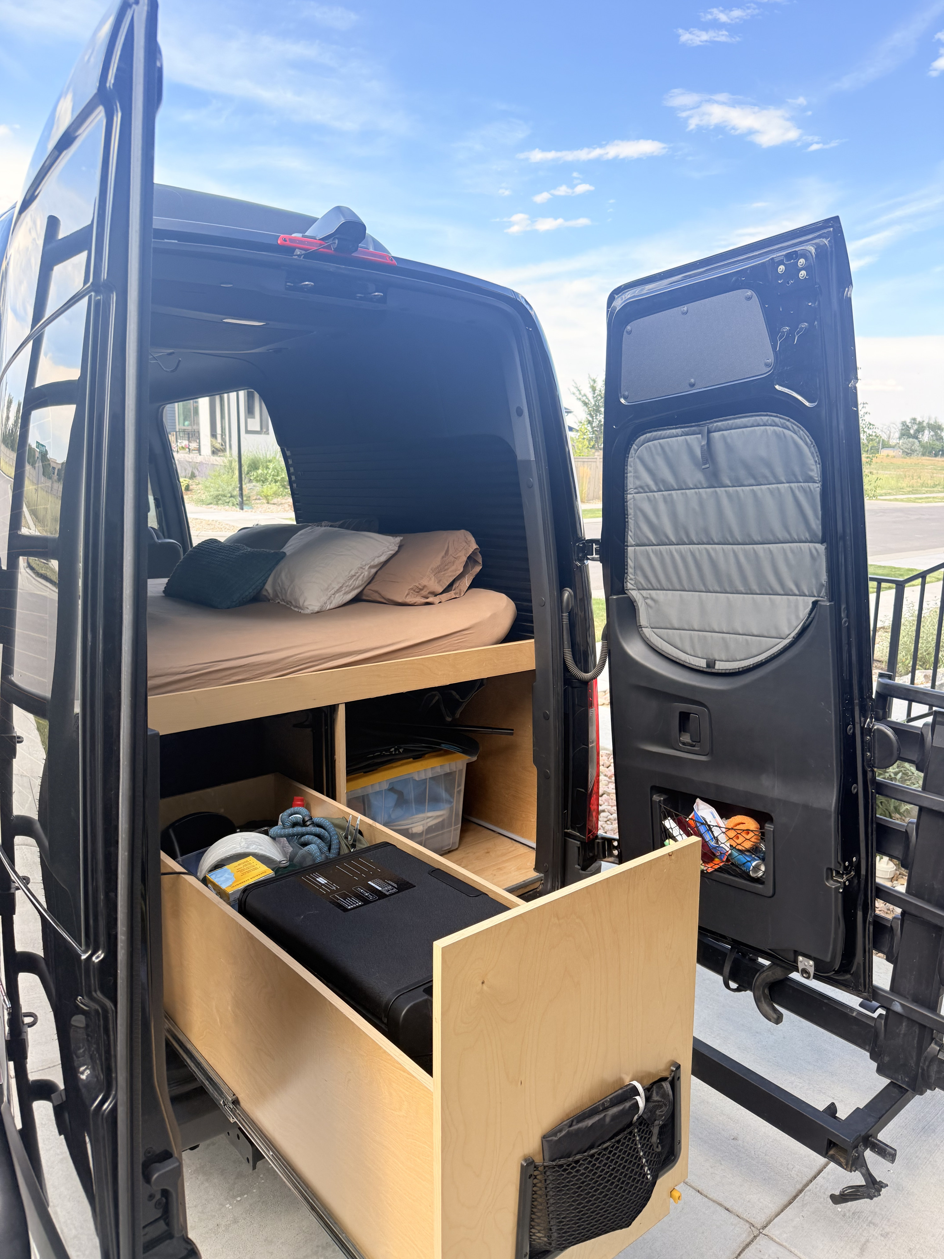 Rear view of an RV rental van's interior, showcasing a bed with pillows and an accessible pull-out storage drawer system.. Mercedez Benz Sprinter 2023