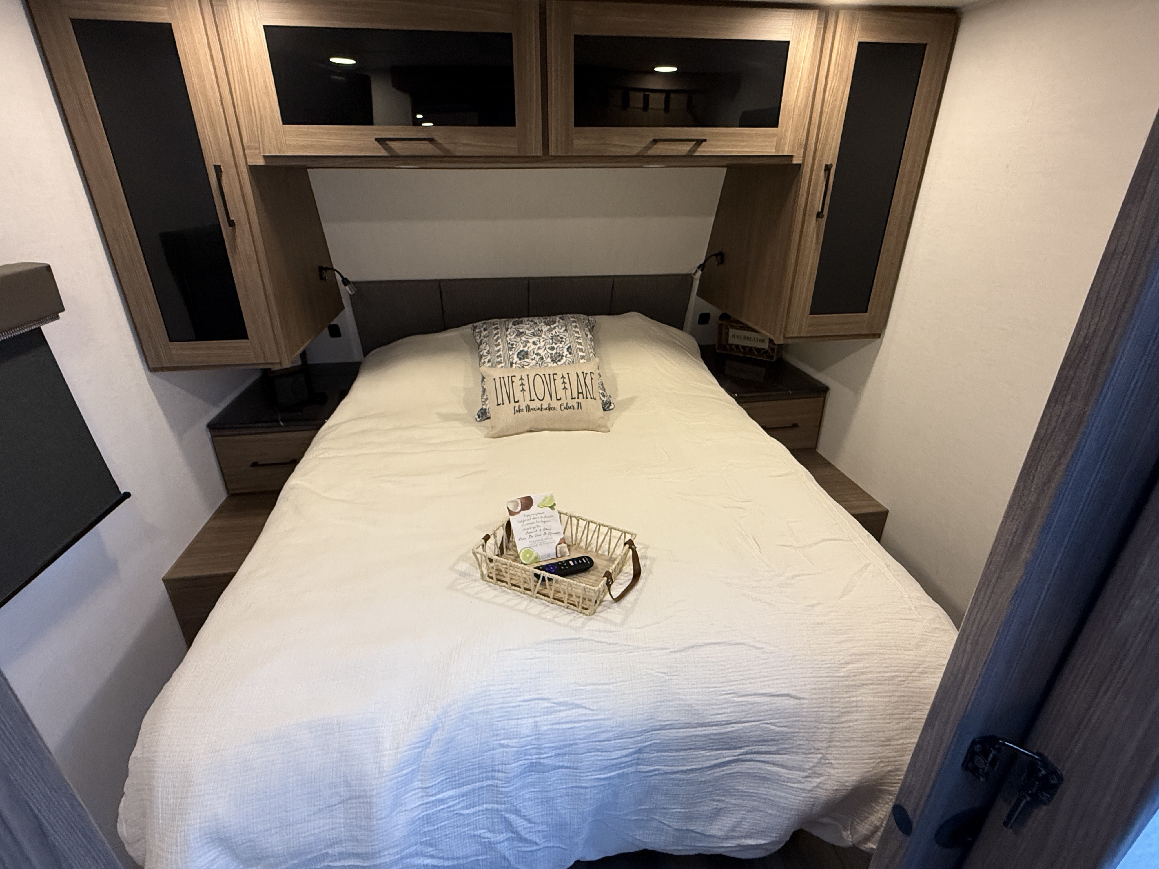 Cozy RV rental bedroom with a queen bed, crisp white bedding, overhead cabinets, and nightstands. A welcome basket awaits.. Alliance RV Delta 2025