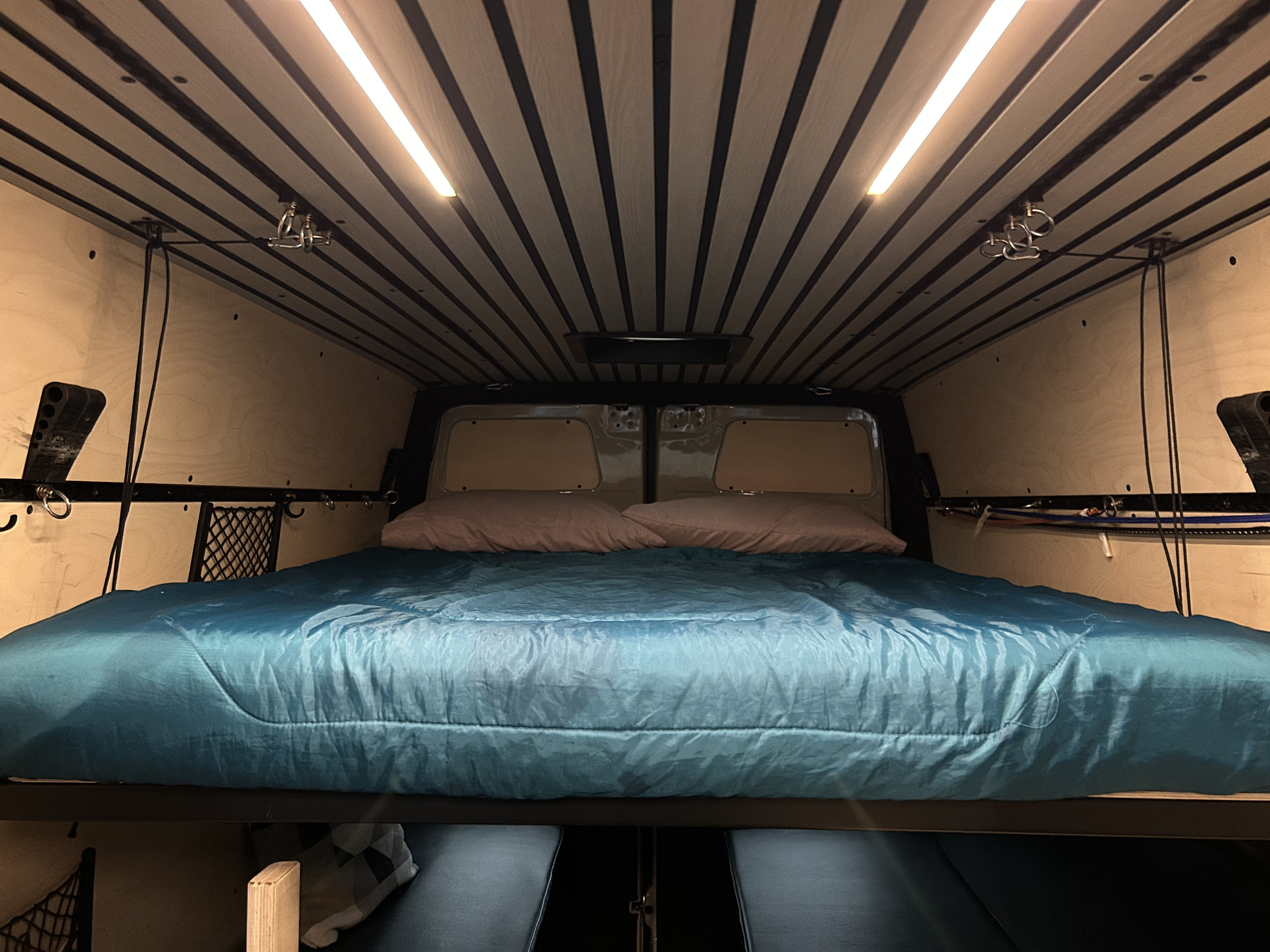 RV rental camper van bedroom: a bed with blue comforter, wood-paneled walls, and bright slatted ceiling lights.. Mercedes-Benz Sprinter 2024