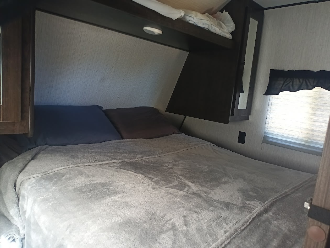 Compact RV rental bedroom featuring a comfy bed, pillows, overhead cabinets, and a window with blinds.. Heartland RVs Prowler 2021