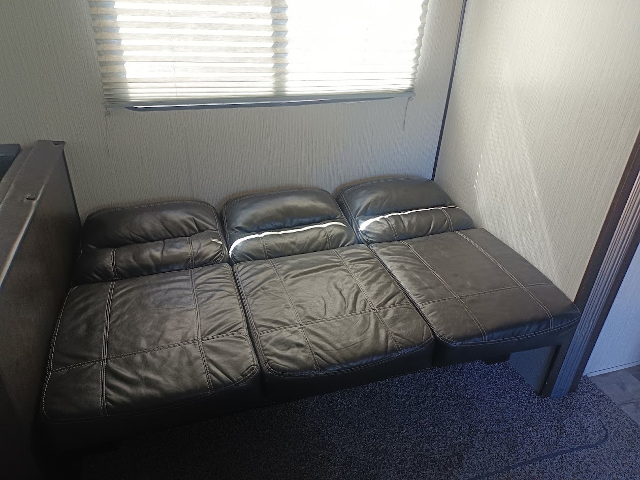 Dark faux leather sofa, likely a sleeper, in an RV rental. Features a window with blinds above and carpeted floor.. Heartland RVs Prowler 2021