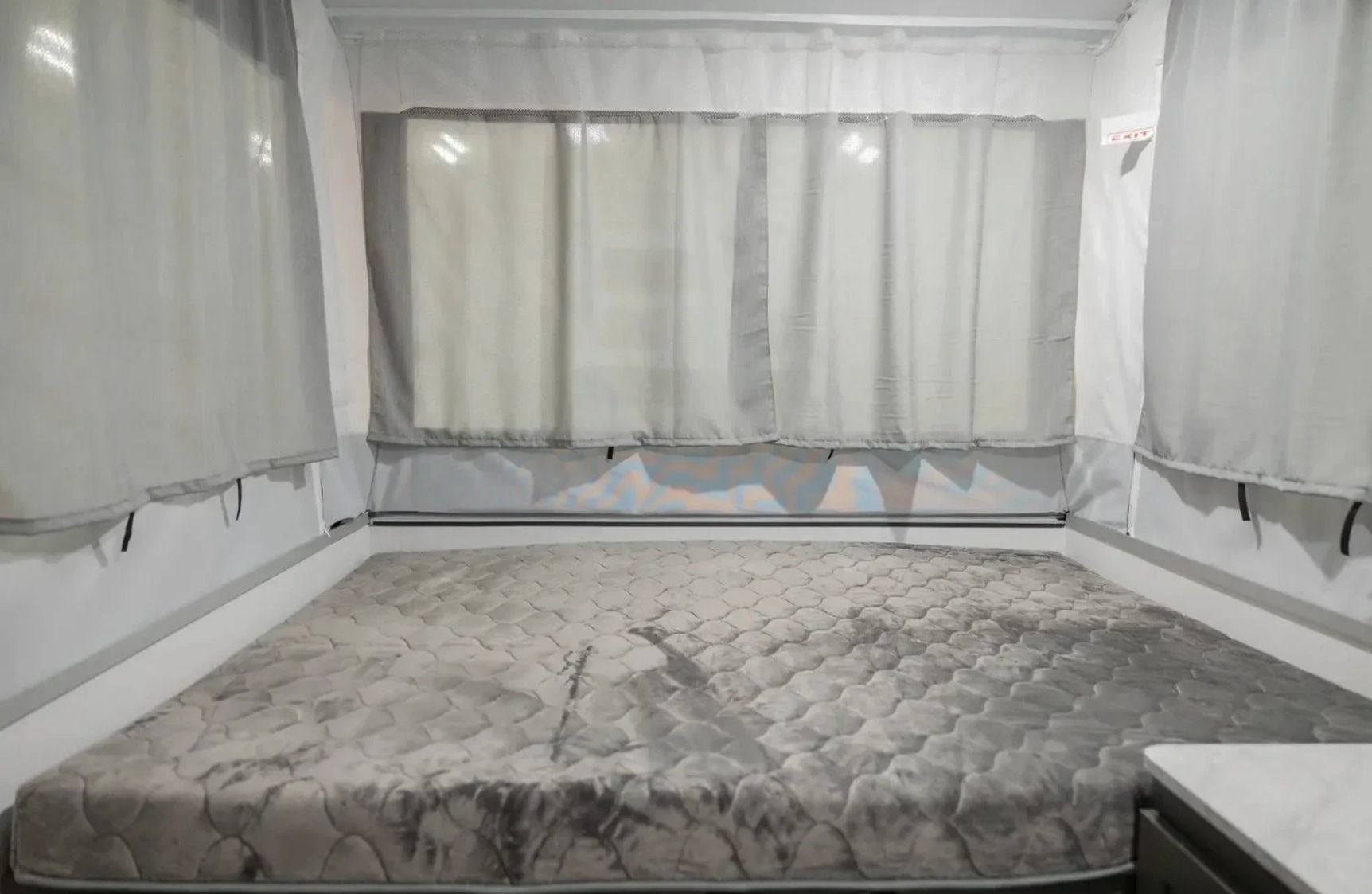 Cozy RV rental pop-up camper bed with a quilted mattress & sheer curtains, an exit sign visible on the right.. Forest River OTG 2025