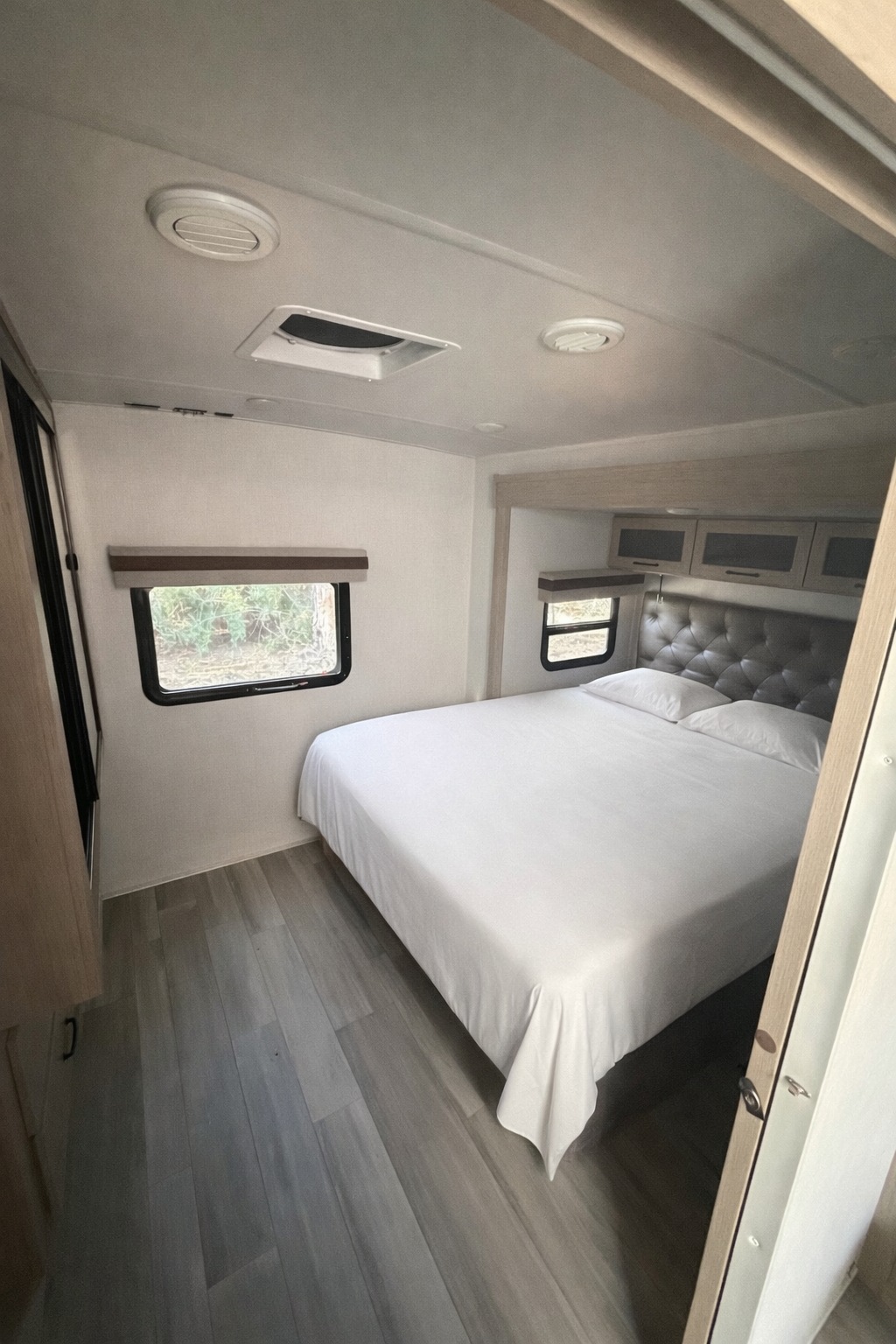 Modern rv rental bedroom features a queen bed with a tufted headboard, overhead cabinets, two windows, and wood-look flooring.. Forest River Rockwood Mini Lite 2024