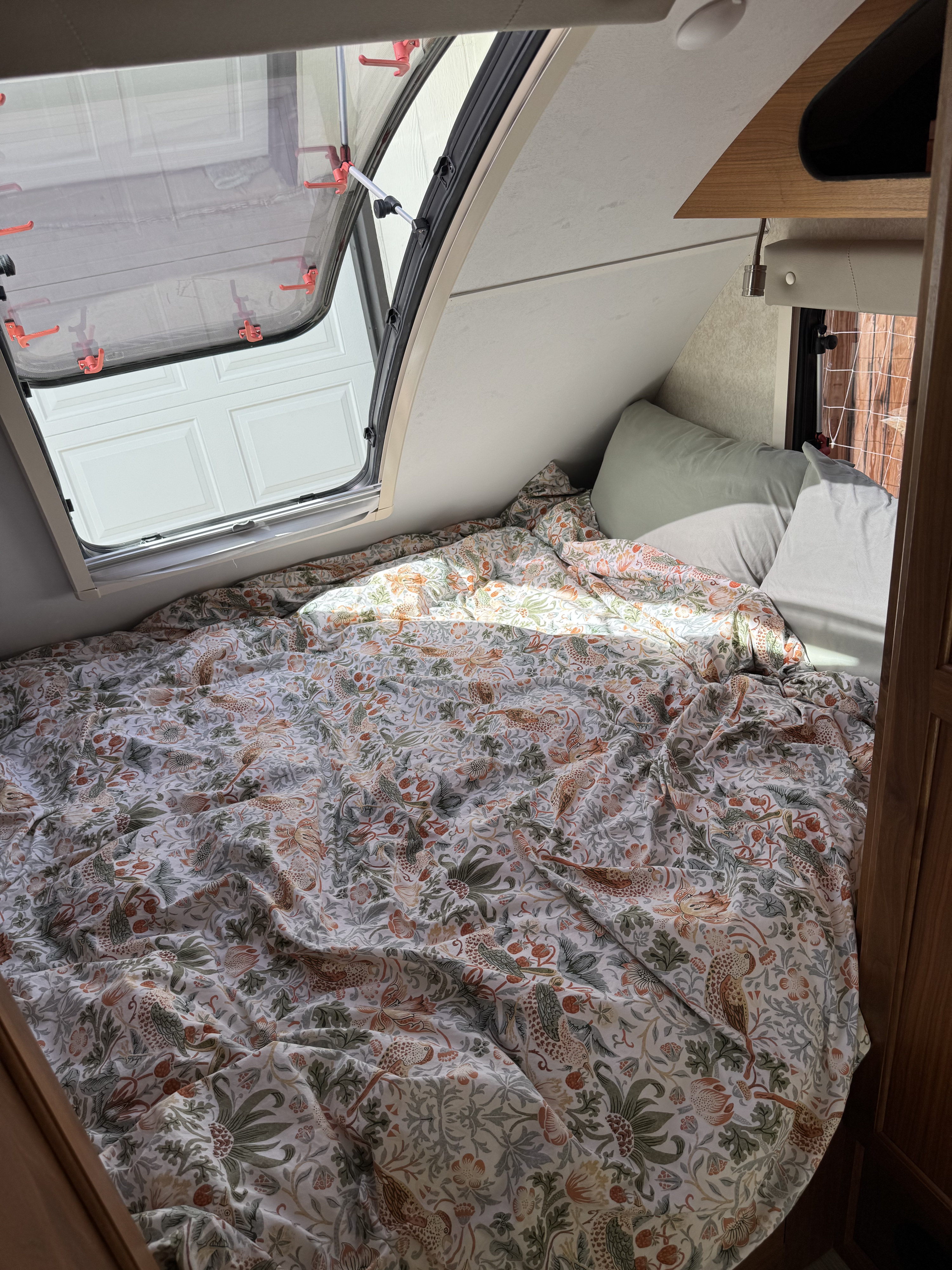 Inside a cozy RV rental: a bed with a detailed bird & floral duvet, light pillows, and an open window showing a garage.. Little Guy Little Guy Trailer 2016