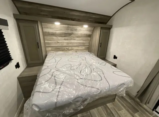 This RV rental bedroom features a bed with plastic-covered mattress, headboard with lights, and side storage.. Gulf Stream Trailmaster 2022