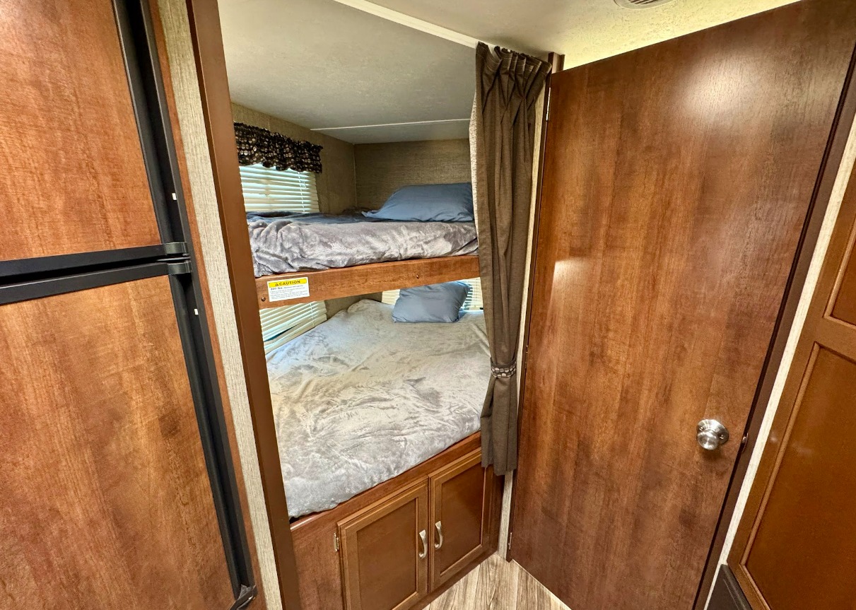 This RV rental interior features neatly made bunk beds with grey bedding and blue pillows. A fridge and wood-paneled cabinets are visible.. Keystone Passport 2017