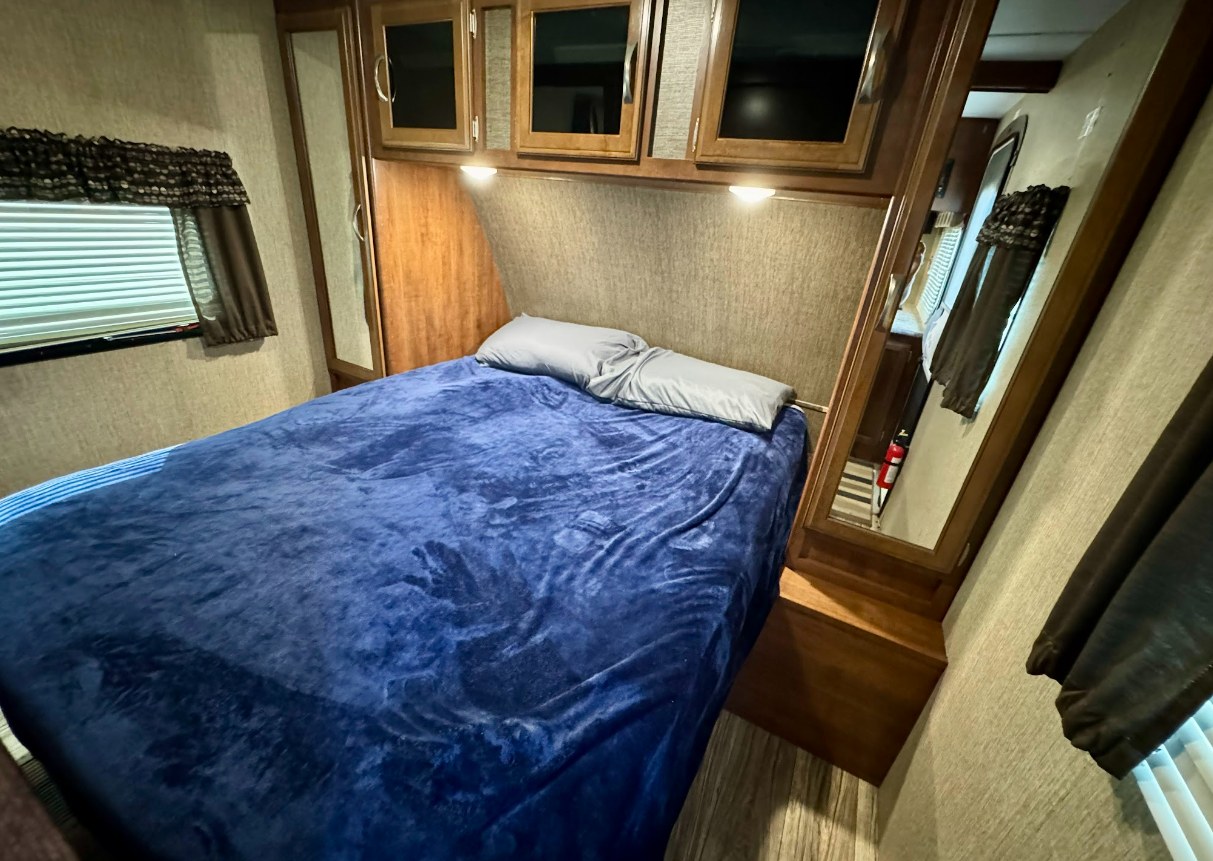 Cozy RV rental bedroom featuring a bed with a blue blanket, overhead cabinets, and a mirror, perfect for travelers.. Keystone Passport 2017