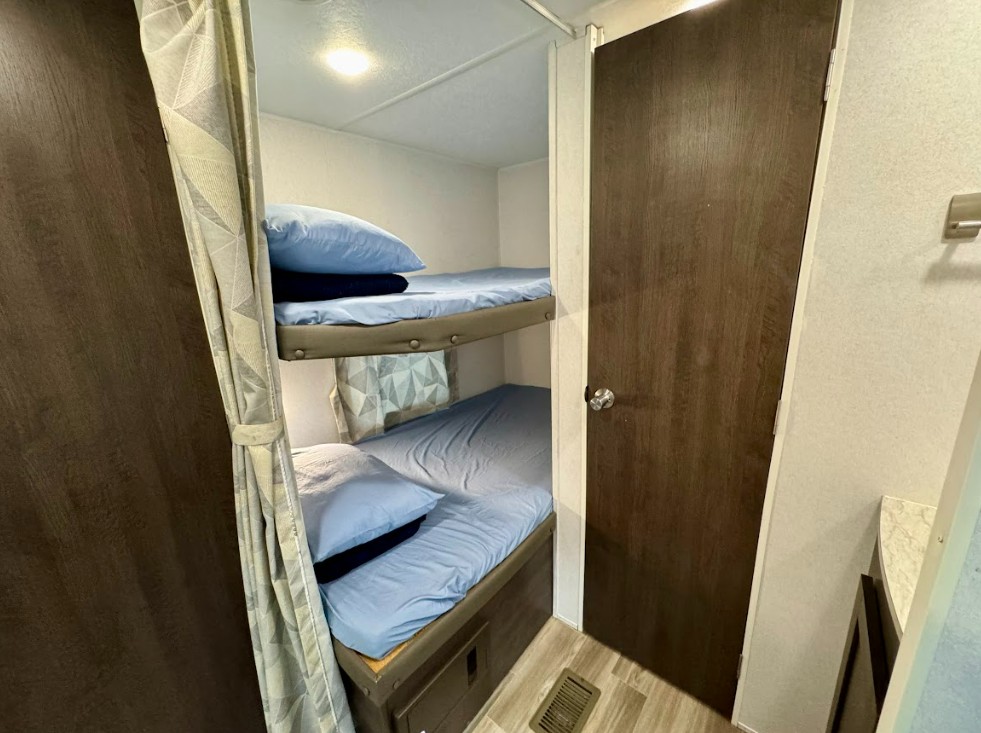 Cozy bunk beds with blue sheets and pillows are visible in this RV rental interior, alongside a privacy curtain and a closed door on the right.. Salem 26DBUD 2020