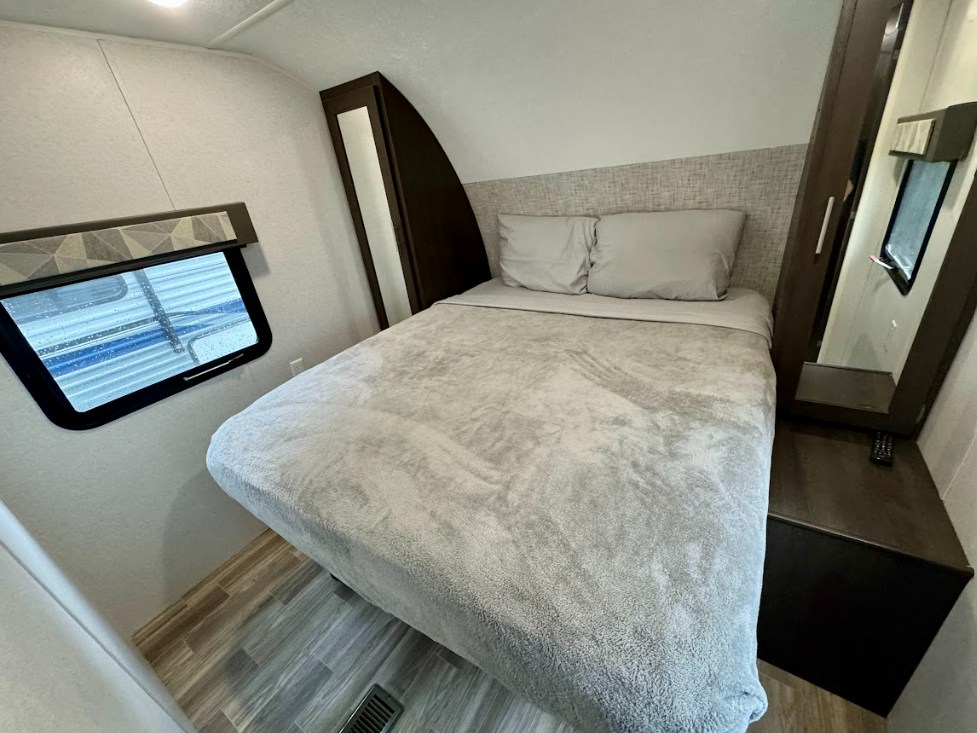 Cozy RV rental bedroom with a plush grey bed, light headboard, window, and dark wood storage unit.. Salem 26DBUD 2020