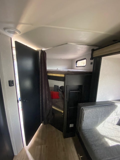 An RV rental interior reveals bunk beds behind a curtain, an open door, and a gray sofa in a bright, compact space.. Forest River Cherokee Grey Wolf 2022