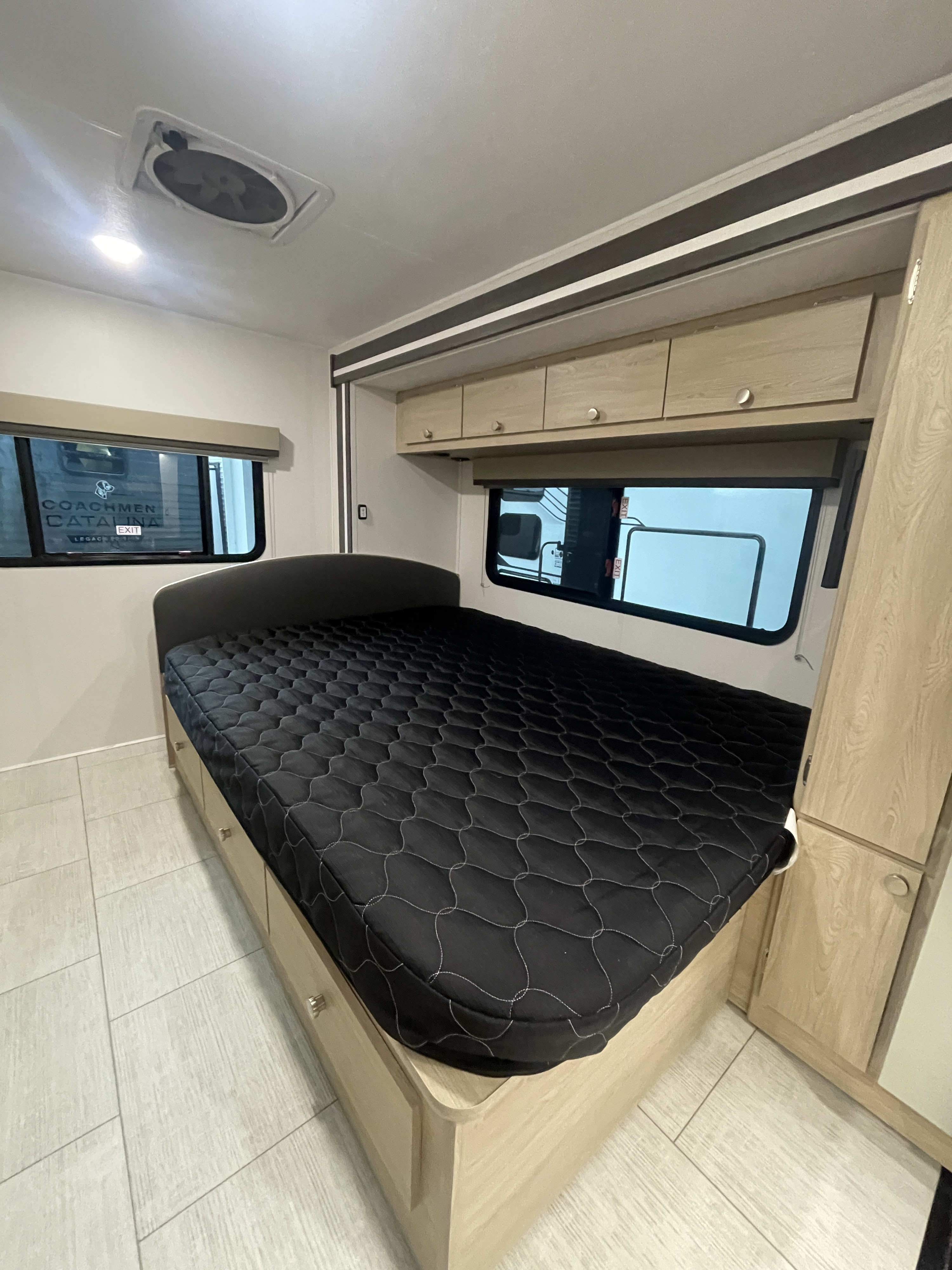 Cozy RV rental bedroom with a black quilted mattress, under-bed storage, overhead cabinets, windows, and light wood finishes.. Coachmen Leprechaun 2026
