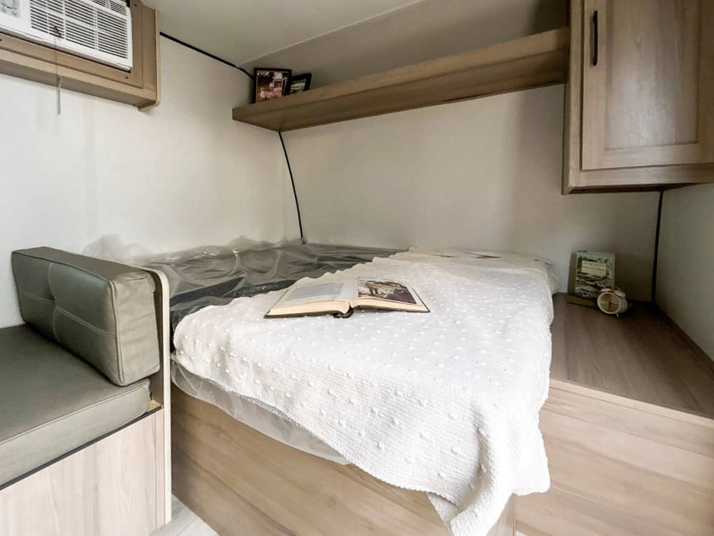RV rental bedroom with a bed, open book, AC unit, and built-in bench seating. A cozy space for travelers.. Keystone Coleman 17B 2026