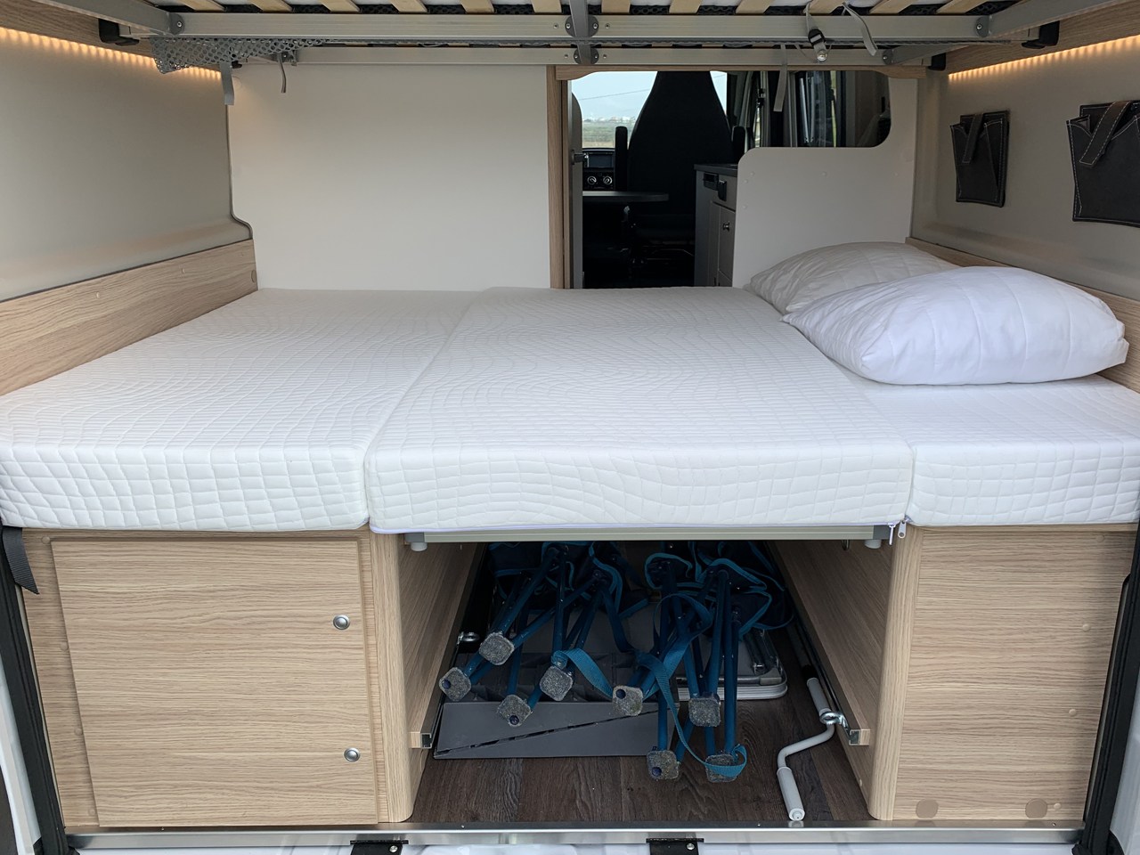 Inside an rv rental: A cozy bed with white mattress over under-bed storage revealing folded chairs and bright lighting.. Sunlight Cliff 600 2023