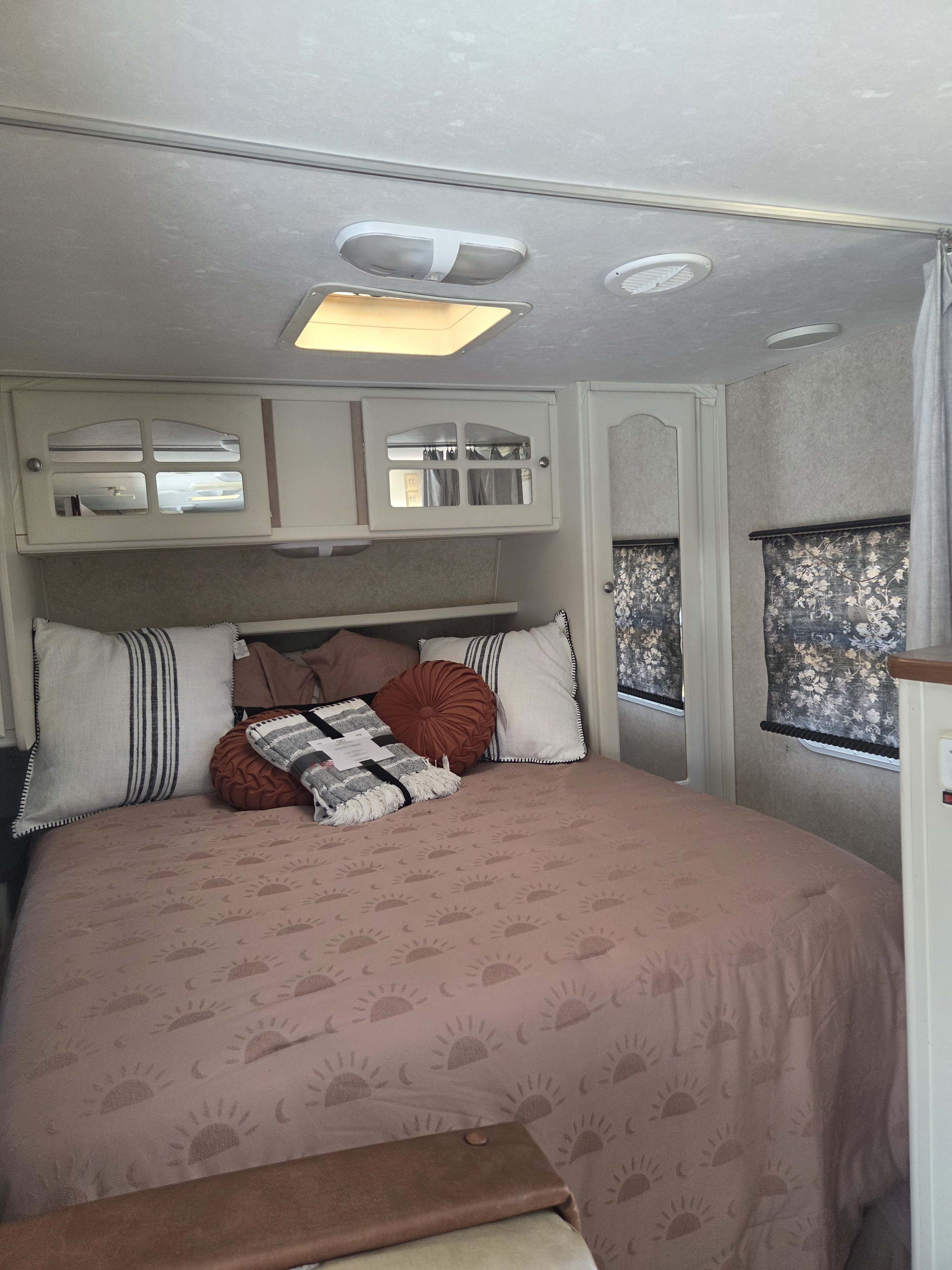 Cozy RV rental bedroom interior featuring a patterned bed, pillows, overhead cabinets, and window curtains for comfortable travel.. Keystone Outback 2006