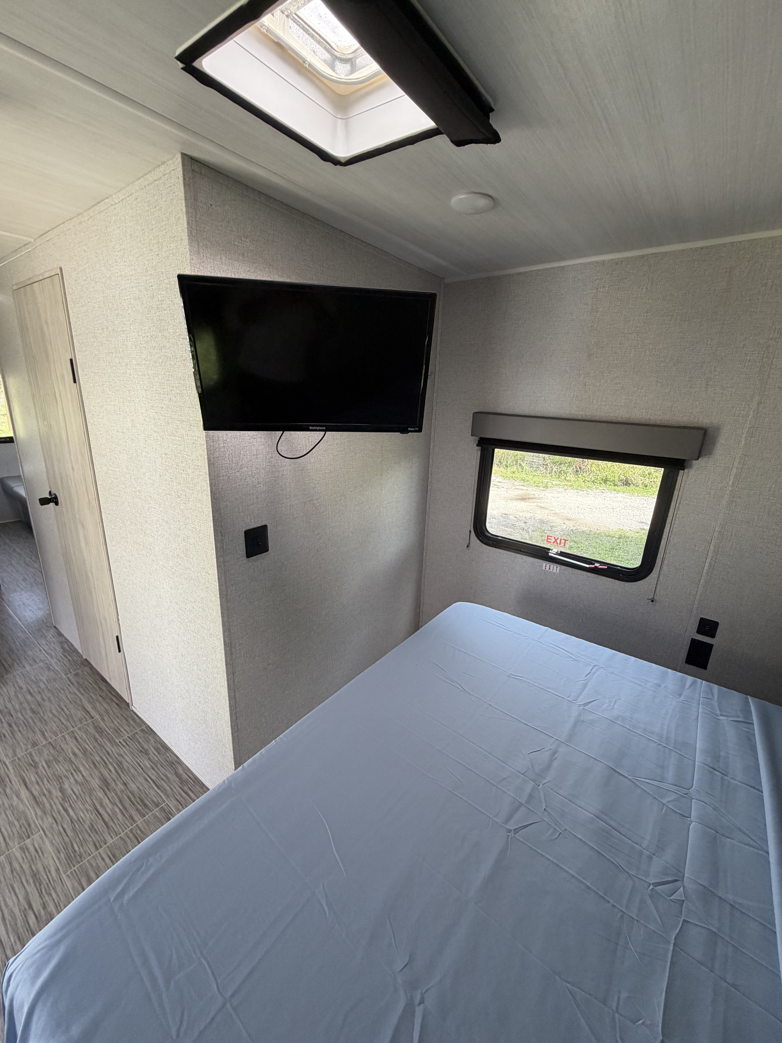 View inside an RV rental bedroom showing a light blue bed, mounted TV, exit window, and ceiling skylight.. East to West Silver Lake 2023