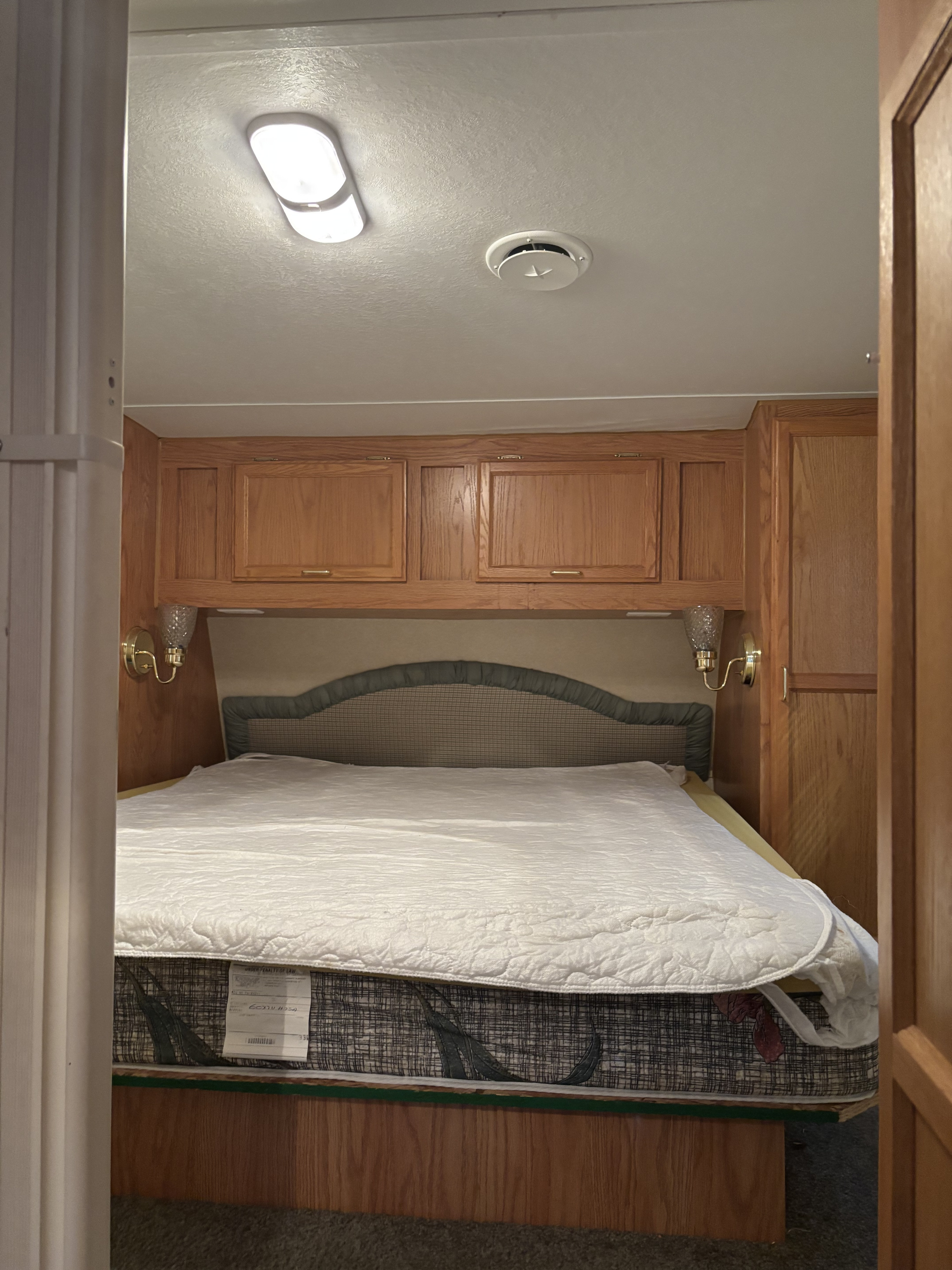 An RV rental bedroom with a bed, patterned headboard, wooden overhead cabinets, and brass wall sconces.. Jayco Qwest 2003