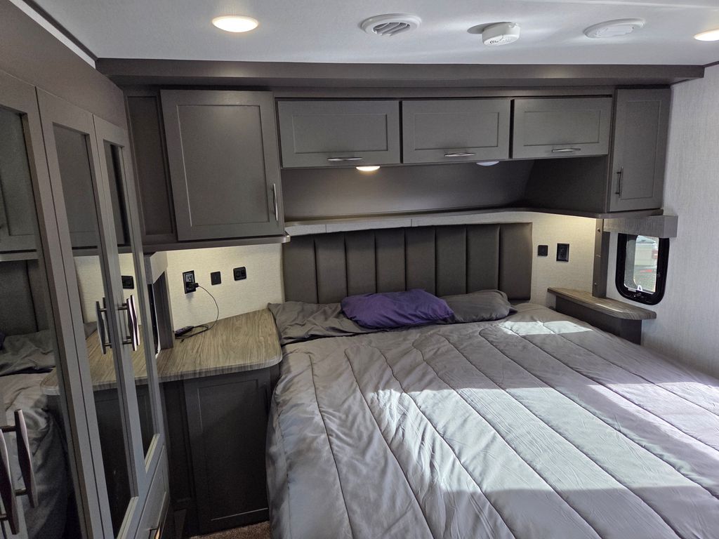 Modern grey RV rental bedroom featuring a queen bed, overhead cabinets, mirrored closet, and practical wall outlets.. Grand Design Momentum Toy Hauler 2023