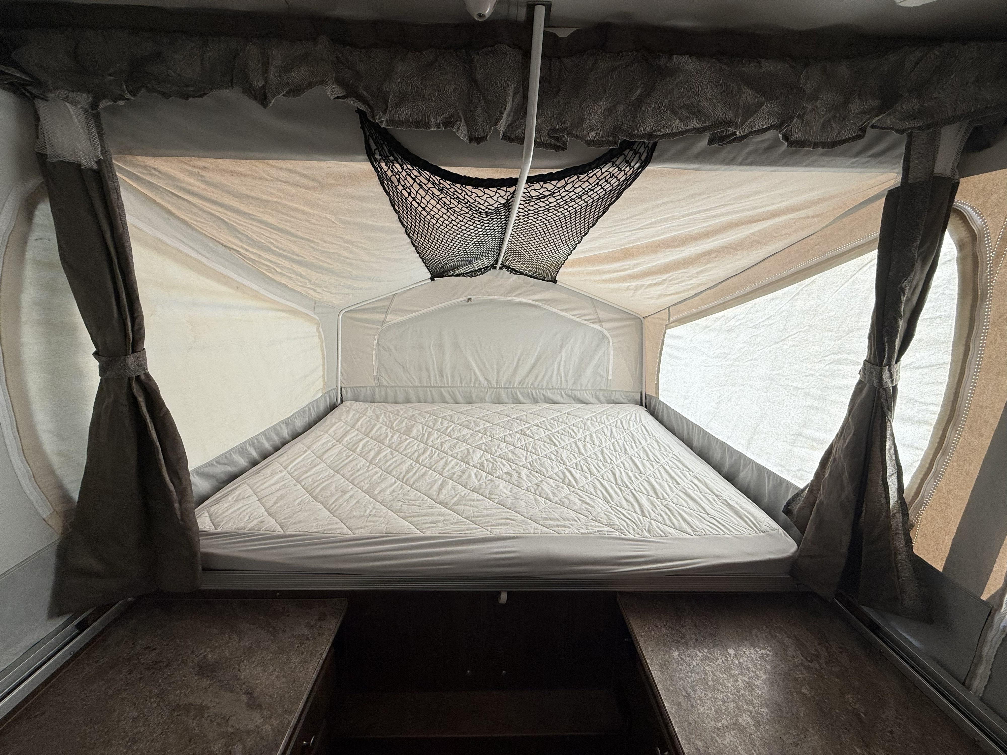 RV rental pop-up camper interior: a comfy quilted bed, light fabric walls, tied curtains, and an overhead mesh storage net.. Forest River Rockwood ESP 2017