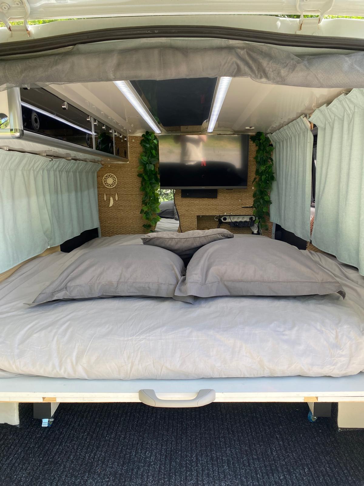 Comfy RV rental bed with a wall-mounted TV, overhead storage, and soft curtains. Your perfect road trip oasis awaits!. Renault Trafic 2018
