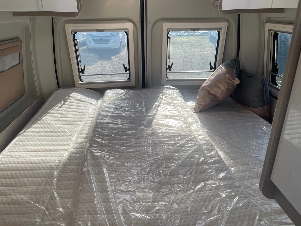 Interior of an RV rental bed area, featuring mattresses still in plastic, two windows with outdoor views, and pillows.. BURSTNER DELFIN C621 LIMITED 2023