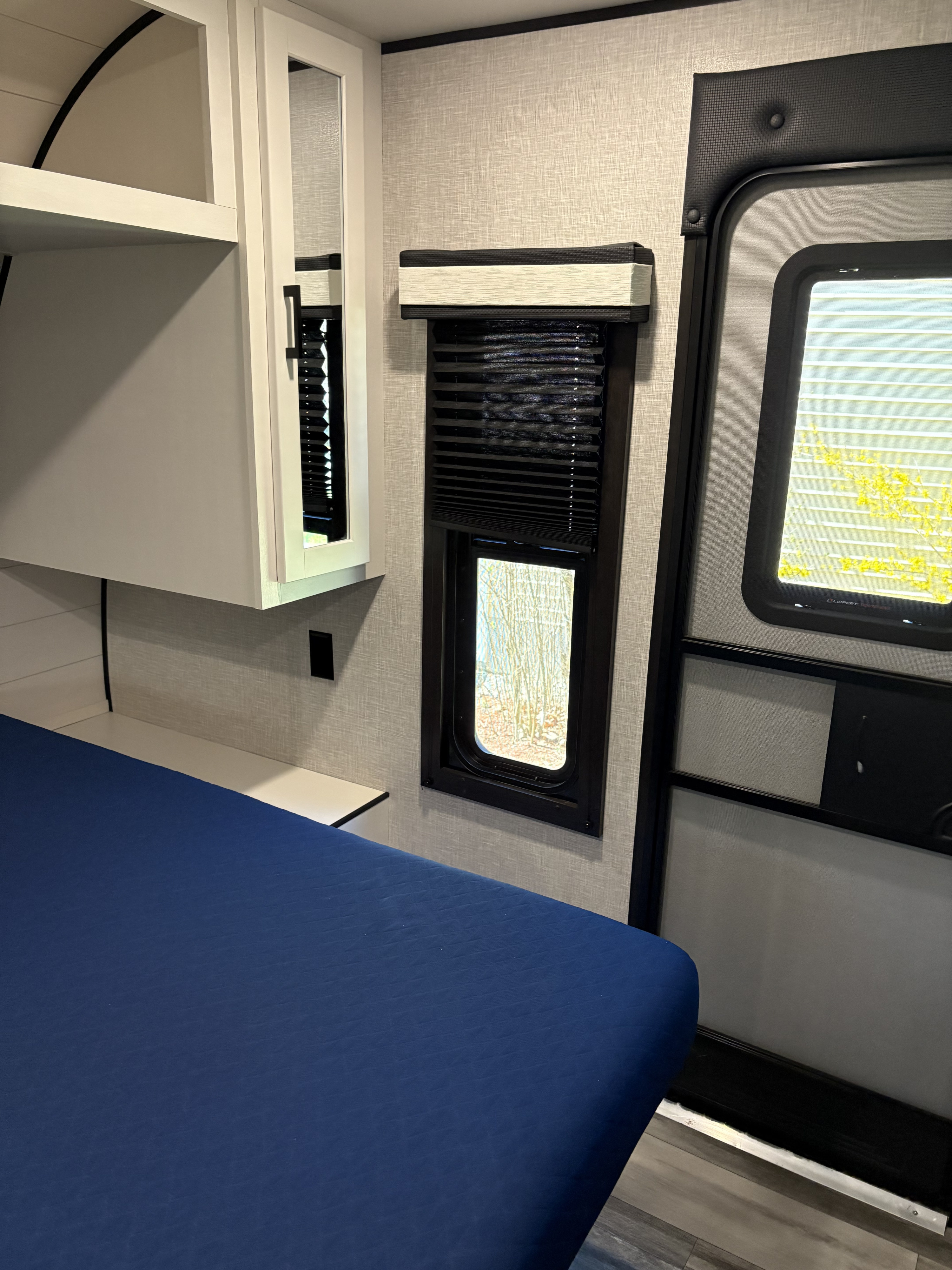 Cozy RV rental bedroom interior featuring a blue bed, overhead cabinets, and window with blackout blinds next to the entry door.. Jayco Jay Flight 2024