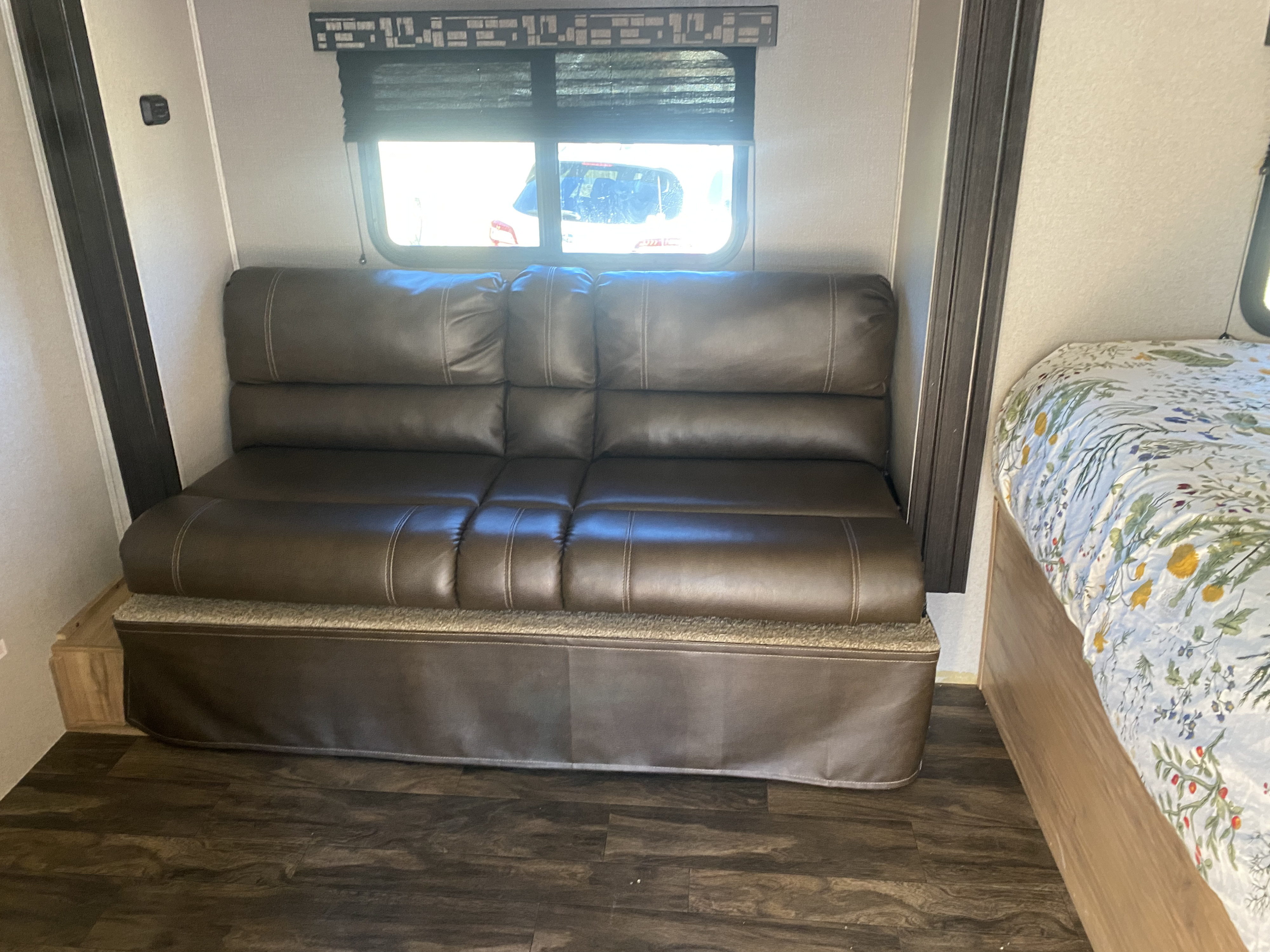 RV rental interior: brown sofa seating, window with blinds, and part of a bed with floral bedding. Modern wood-look floor.. Forest River Coachmen Freedom Express Pilot 2019