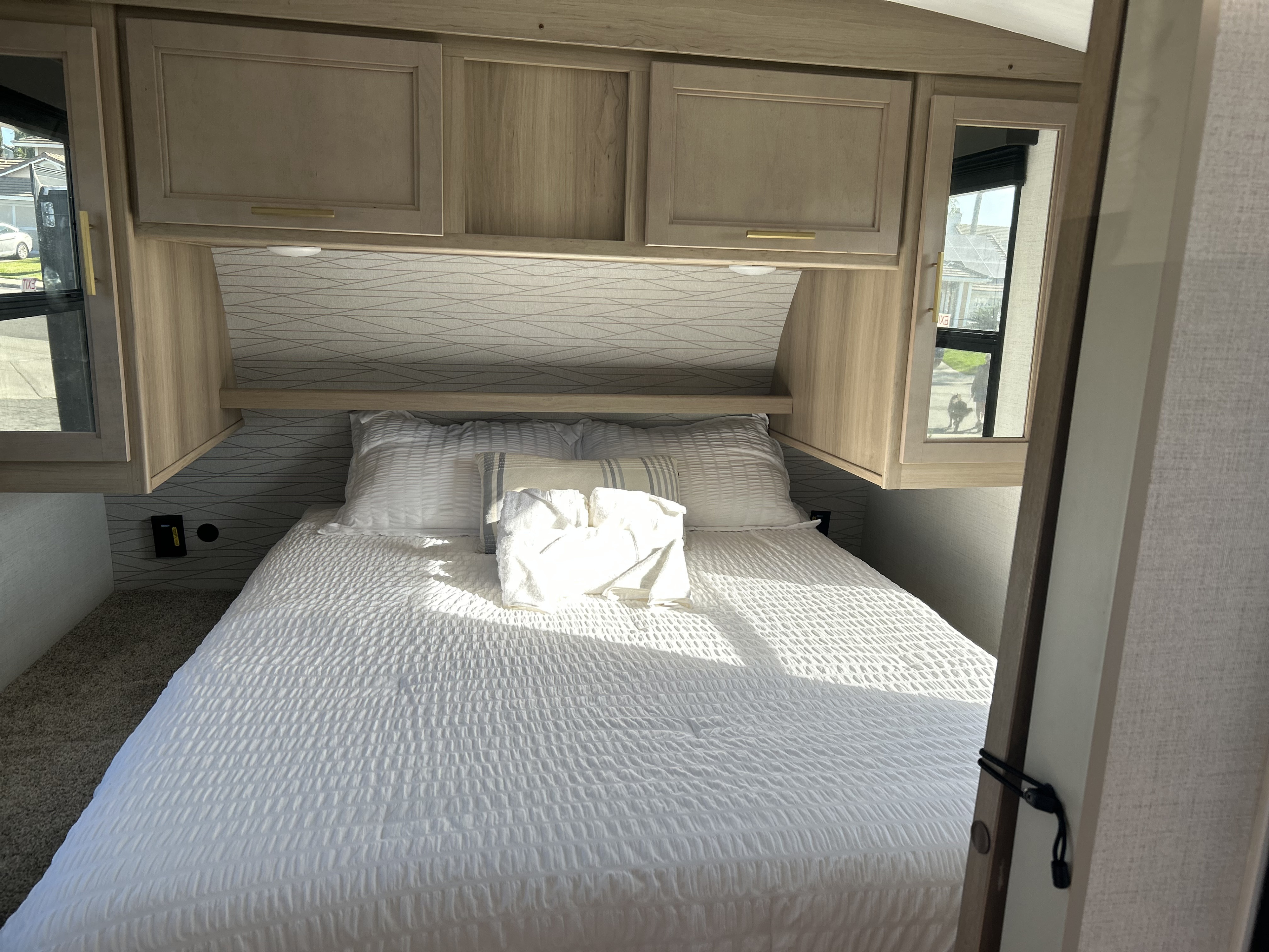 RV rental bedroom: a cozy interior with a white bed, overhead cabinets, patterned wall, and side windows.. Keystone RV Outback Ultra Lite 2024
