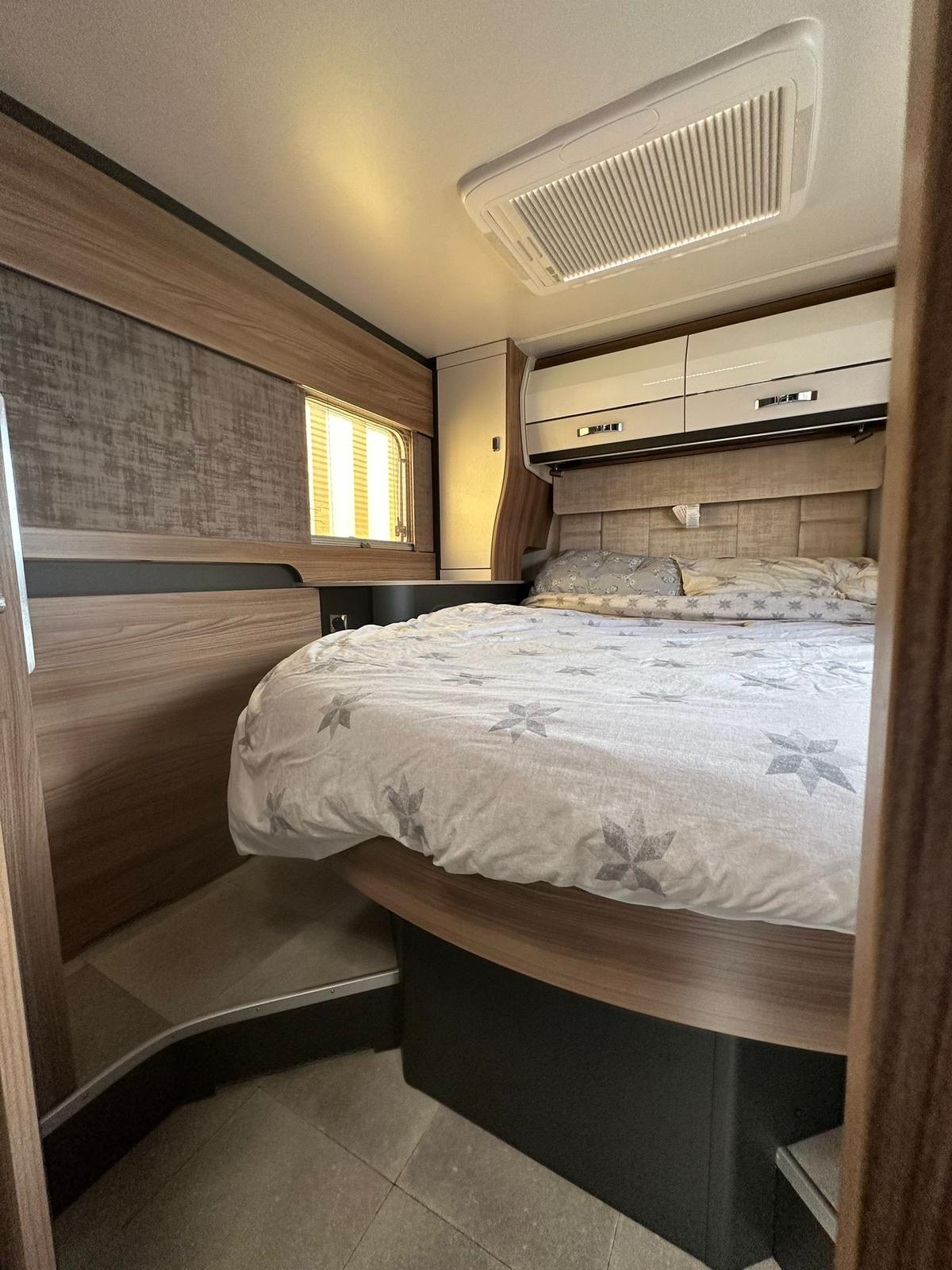 Cozy RV rental bedroom featuring a comfy bed with patterned duvet, overhead storage, AC unit, and a window for natural light.. swift kontiki 2022