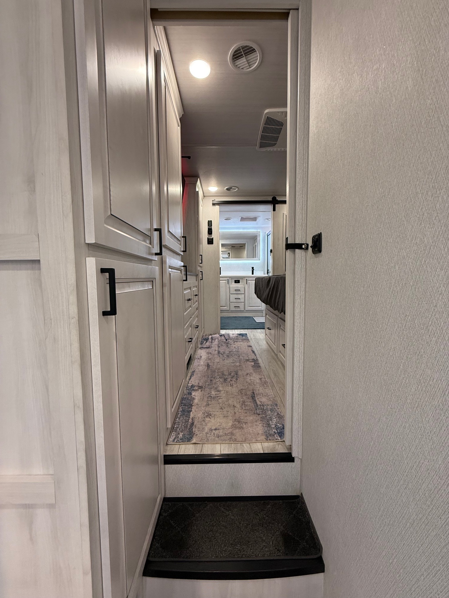 This modern rv rental interior features a bright hallway with light cabinets, leading to a bedroom and bathroom via steps.. East to West Ahara 2024