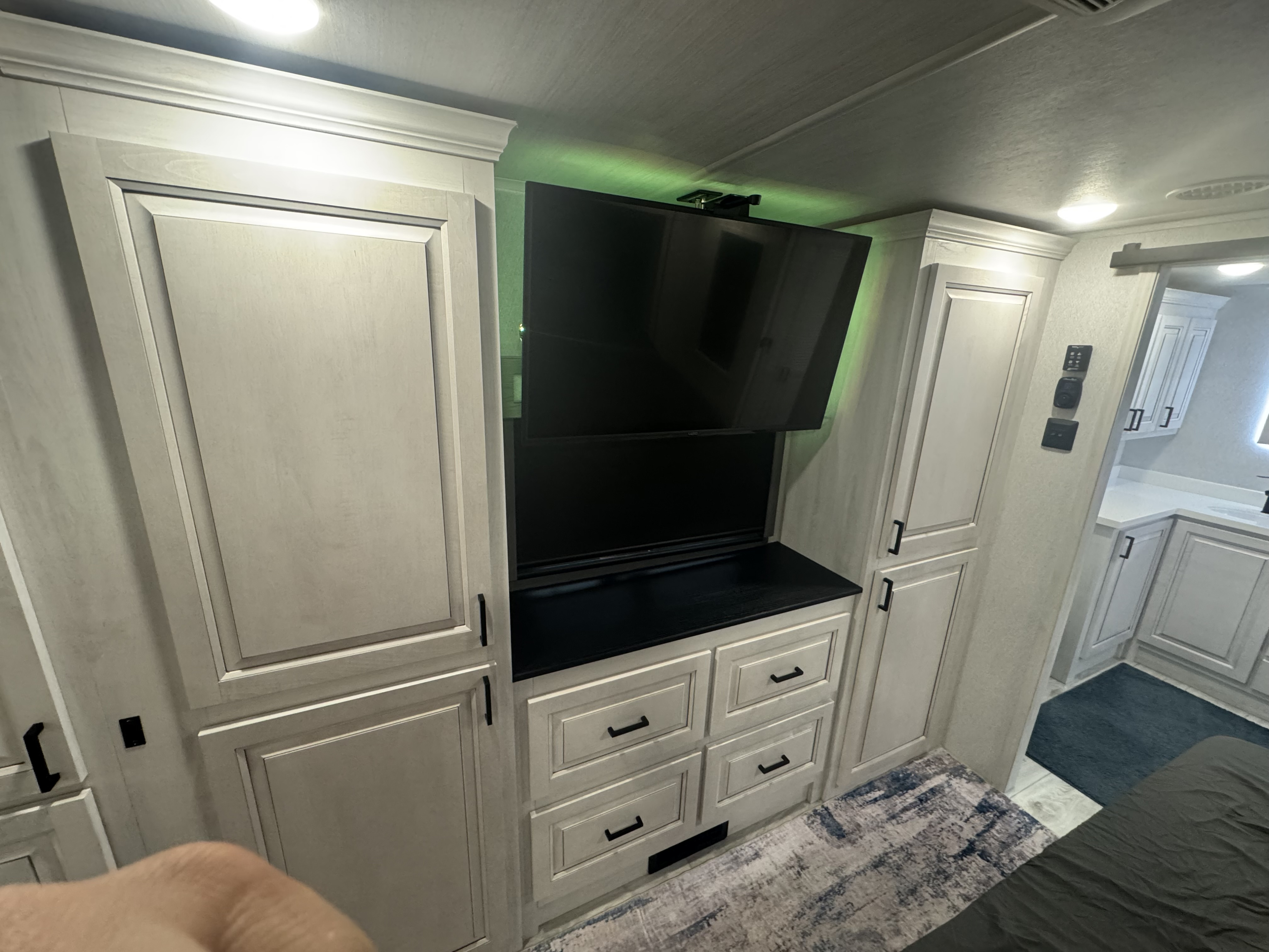 Modern RV rental interior: A bedroom with light wood-grain cabinets, black handles, a mounted TV, and an adjacent vanity area.. East to West Ahara 2024