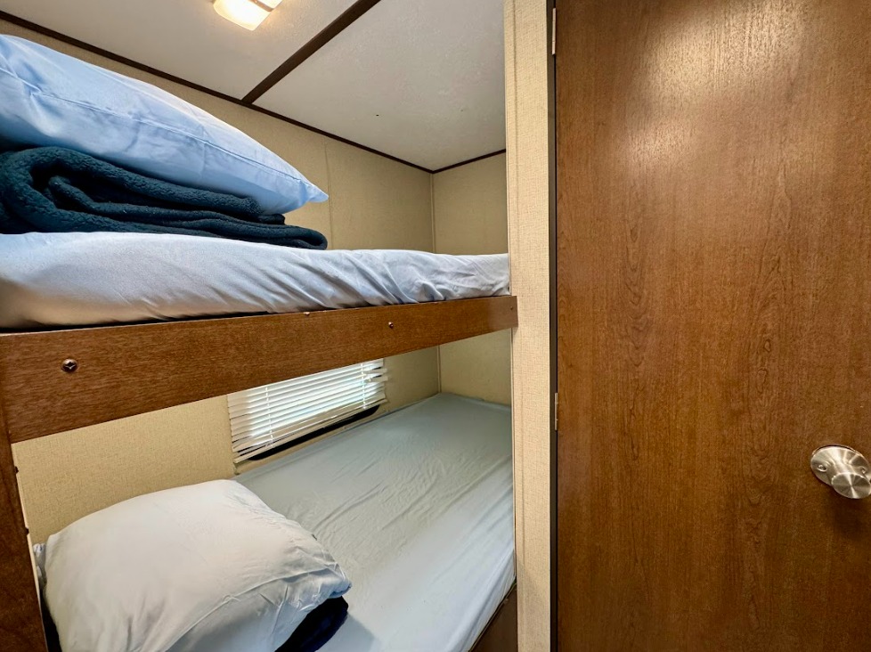 RV rental bunk beds, neatly made with light blue bedding inside a cozy camper. A wooden door is on the right.. Prime Time Avenger 26BH 2020