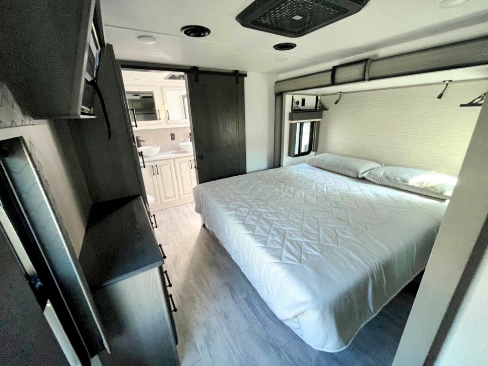 Modern RV rental bedroom: queen bed, wall-mounted TV, dresser, and bathroom visible beyond a sliding barn door.. Keystone RV Montana Legacy 3761 FL 2023