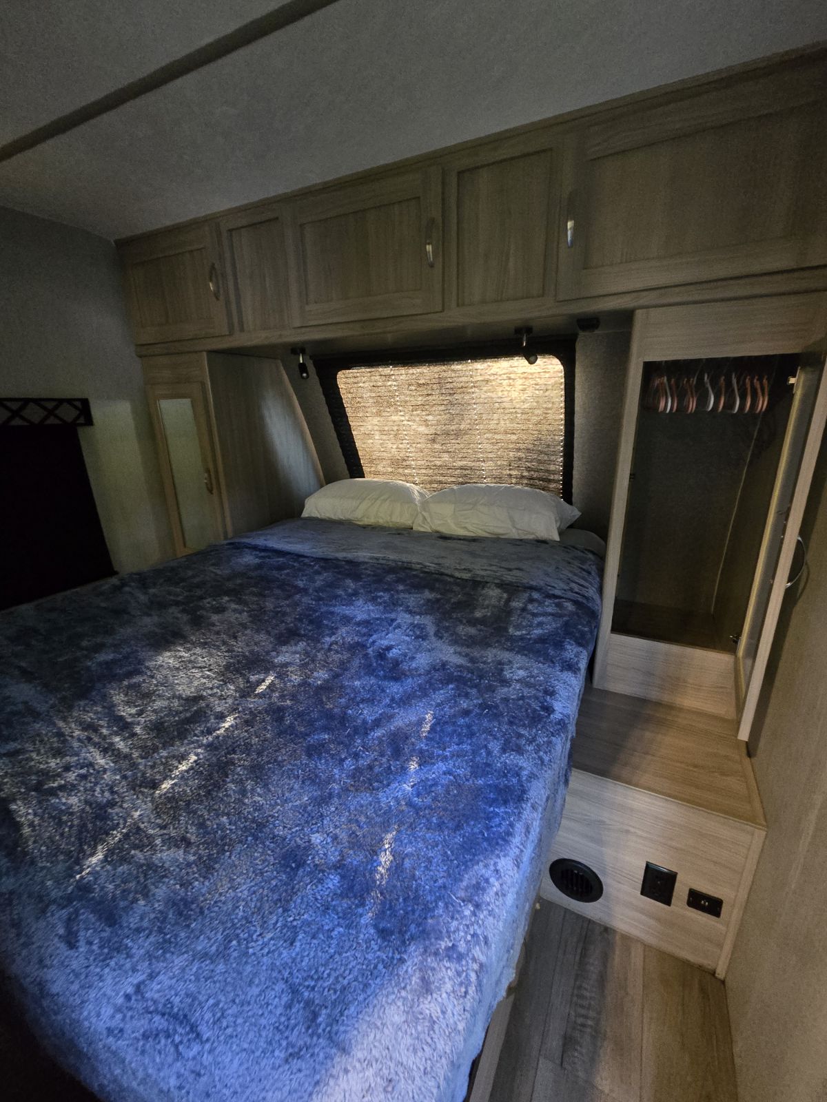 Cozy RV rental bedroom featuring a bed with a blue blanket, overhead cabinets, a window with blinds, and a small closet.. Coachmen Freedom Express Liberty Edition 292BHDSLE 2021