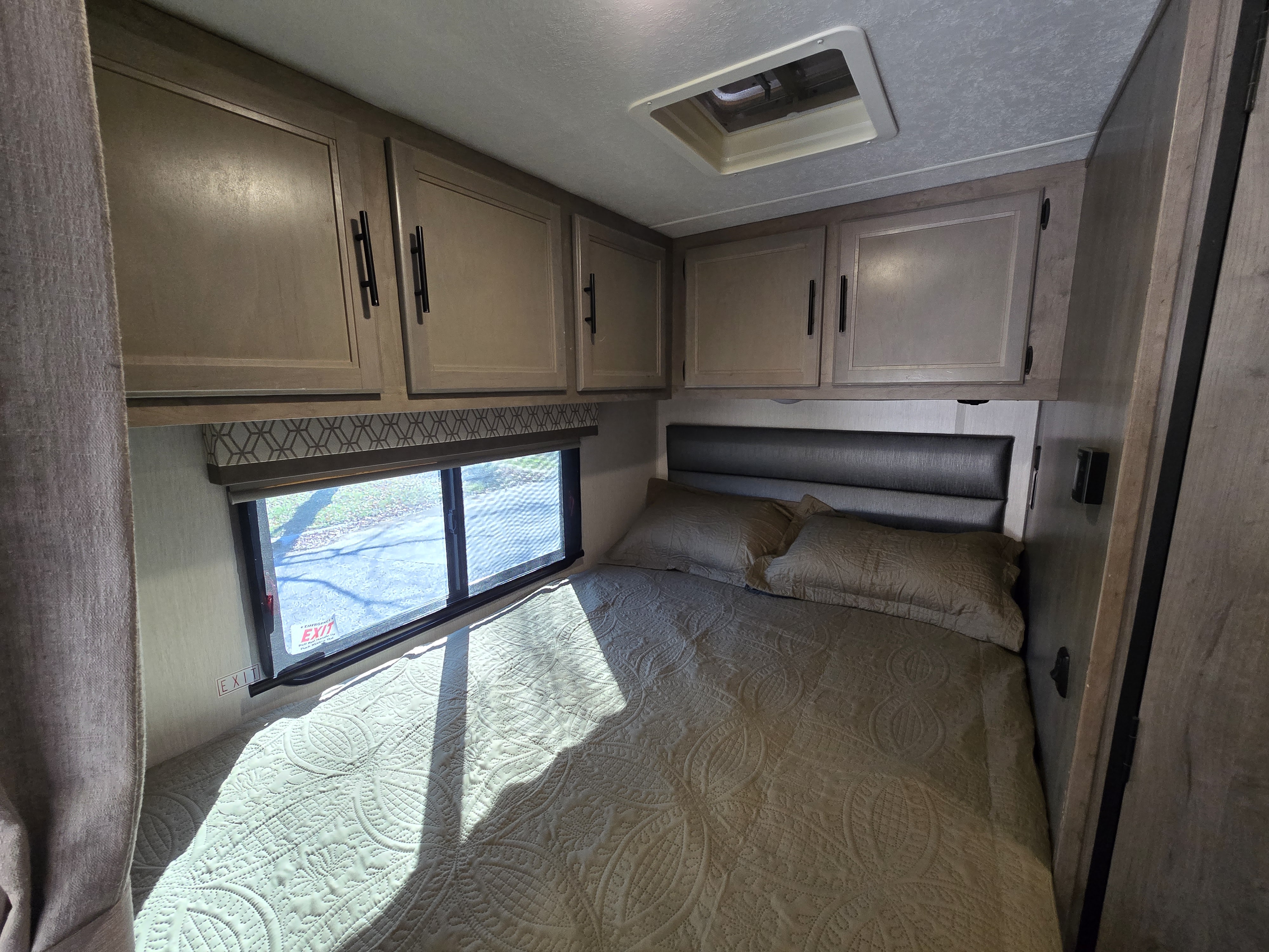 Inside an RV rental: a bright bedroom with a quilted bed, headboard, overhead cabinets, and a window with an emergency exit.. Forest River Coachmen Prism 2020