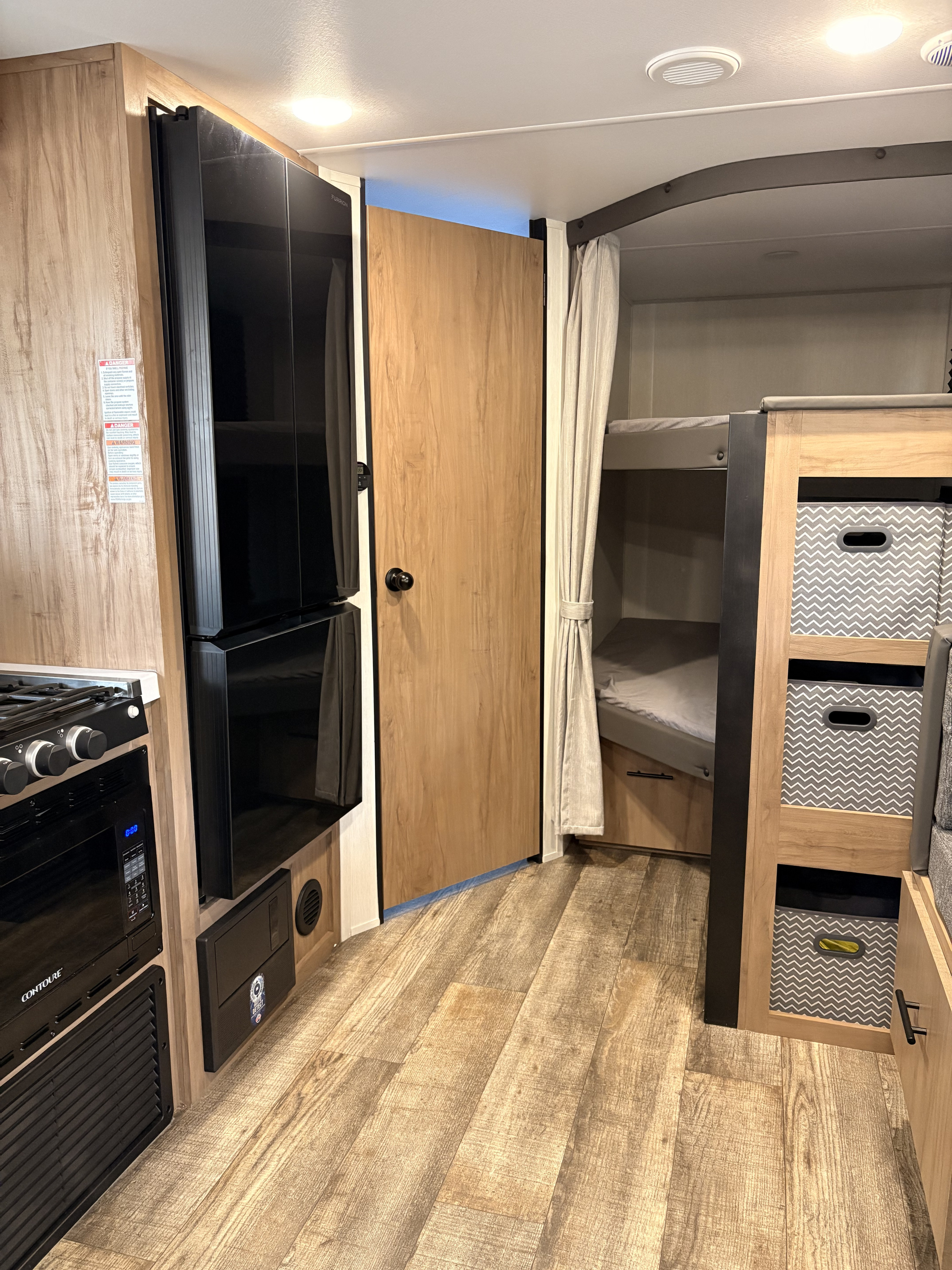 This RV rental interior features a compact kitchenette with a fridge, stove, and microwave, opposite bunk beds with patterned storage bins.. Forest River Campsite Reserve 2024