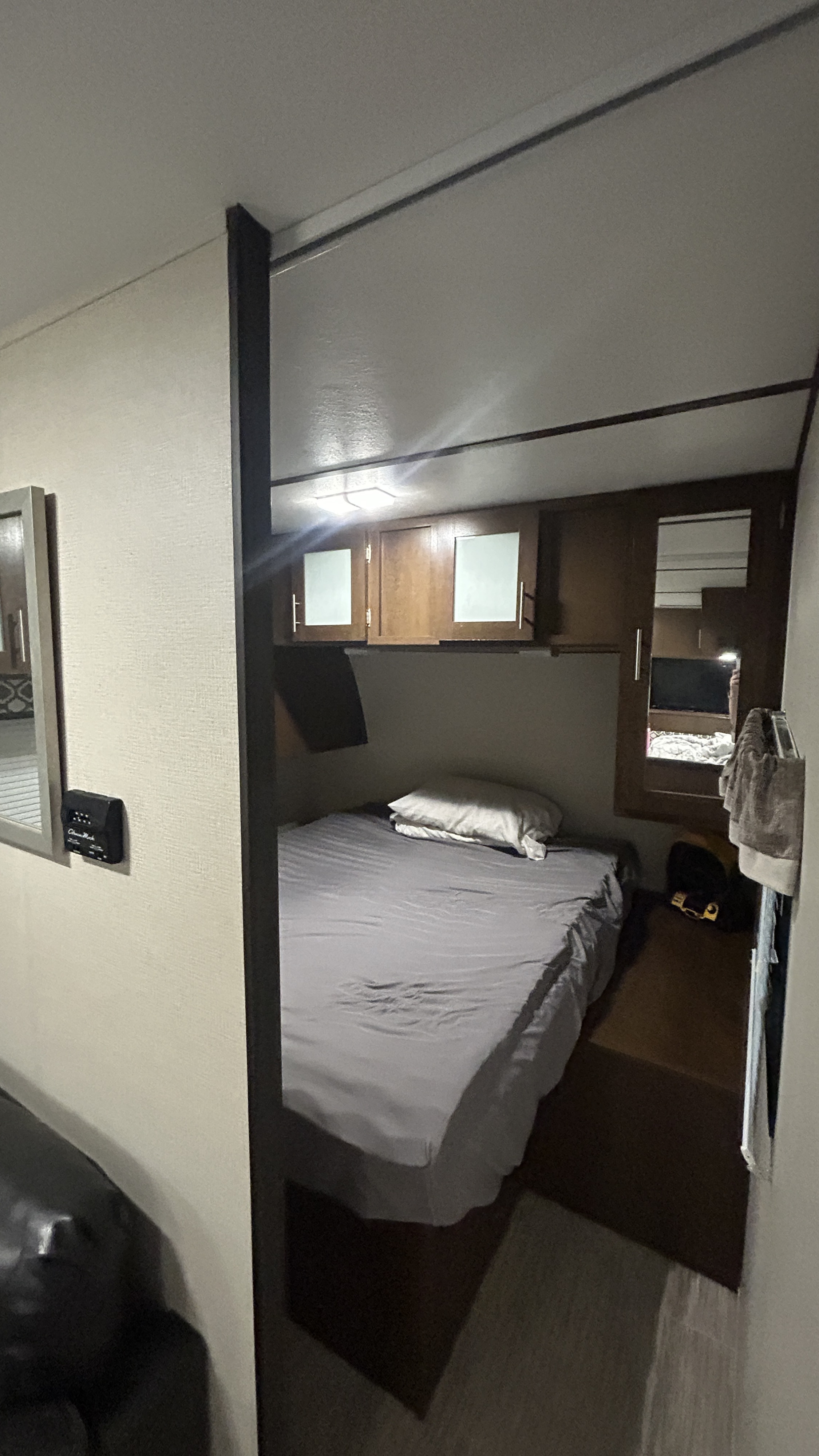 Inside an RV rental, a compact bedroom features a bed with grey bedding, wooden overhead cabinets, and a side mirror reflecting the interior.. Primetime Avenger 2019